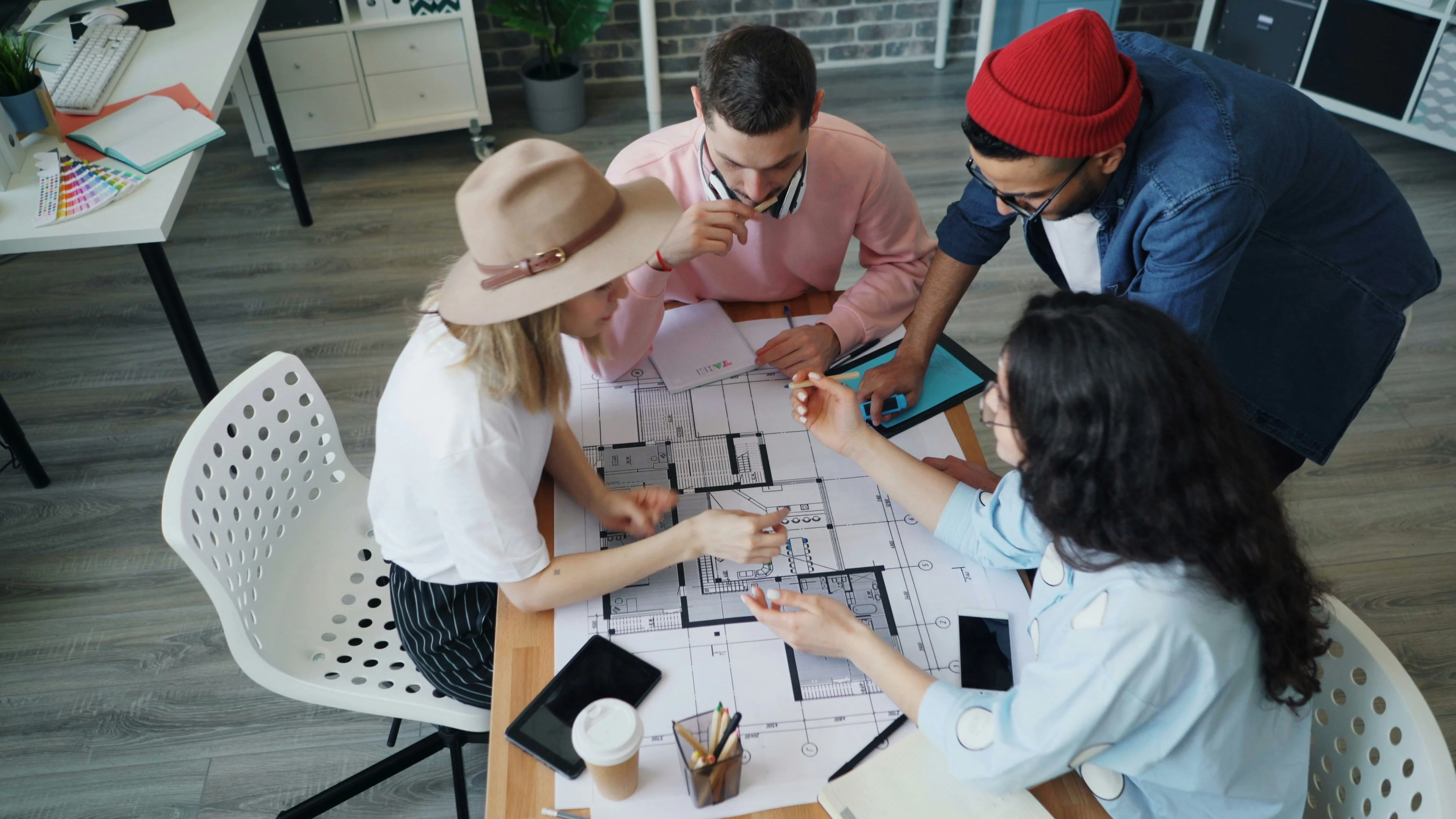 Four people collaborate around a table with architectural plans, discussing and pointing at the designs. Coffee cups and tablets are on the table.