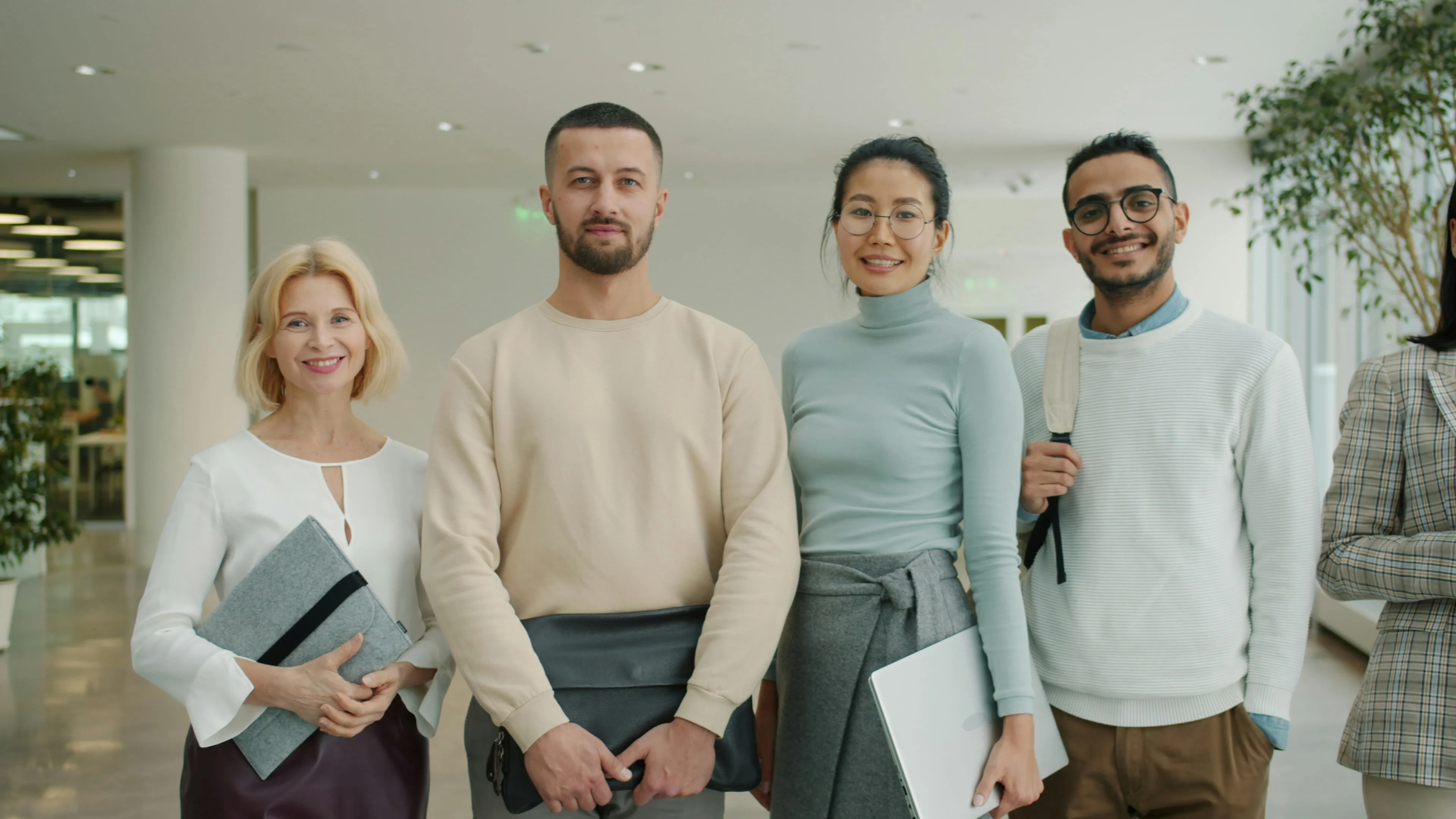 Four people smiling indoors, holding notebooks and folders, standing in a well-lit office space with plants in the background.