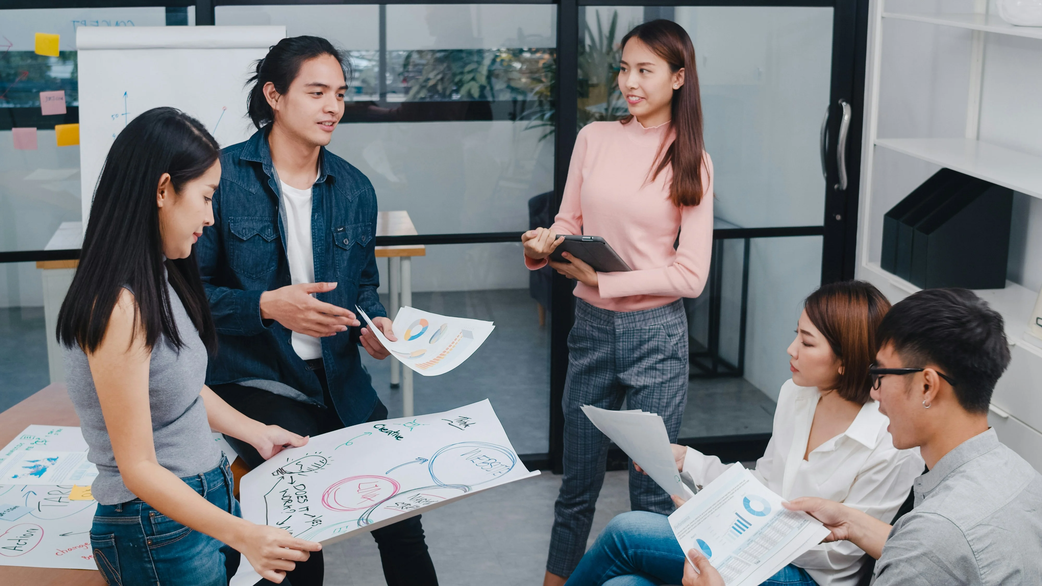Five people in a meeting room discussing charts and graphs. One person stands holding a paper, while others sit and listen attentively.