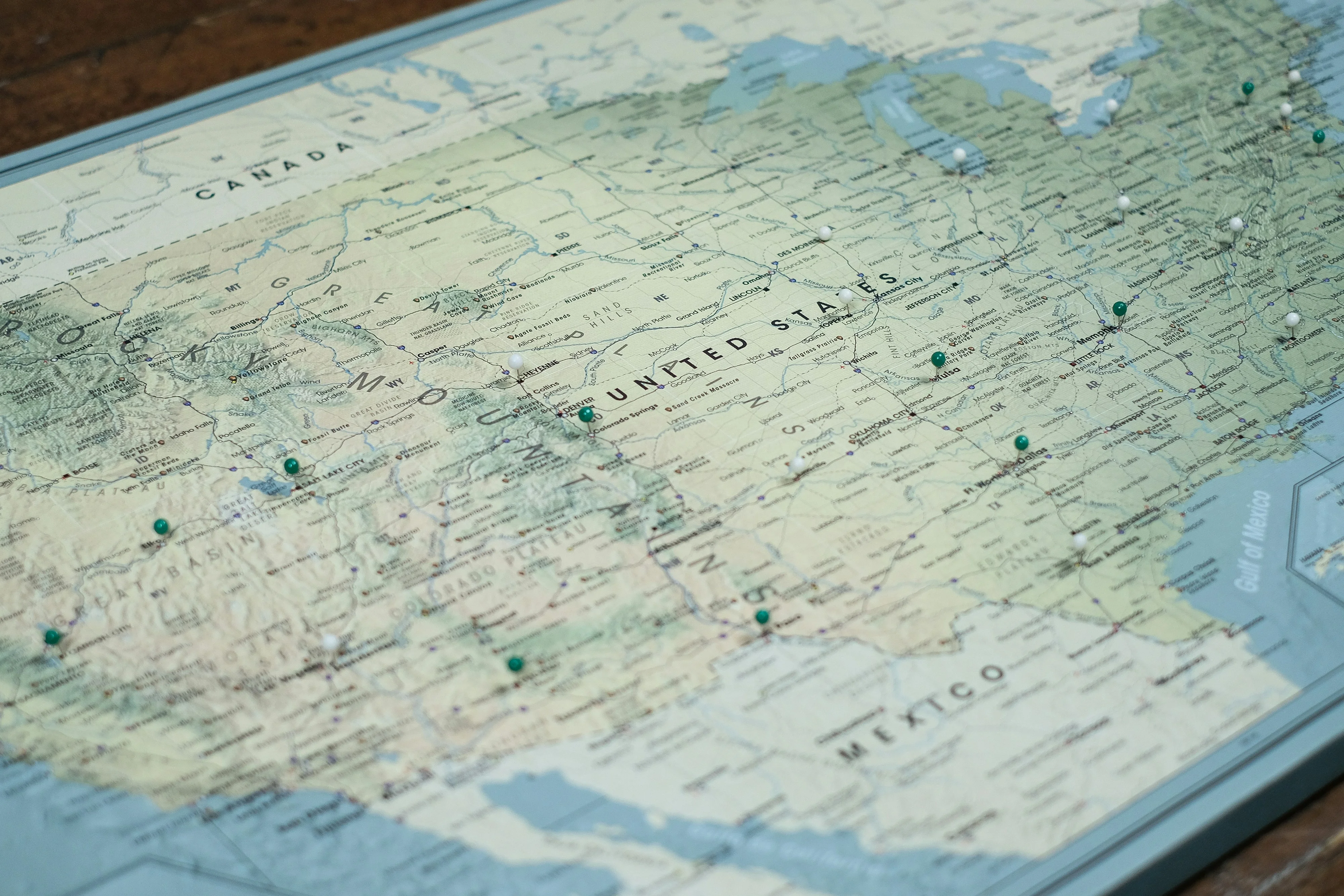 A map of the United States with green pins marking various locations, showing parts of Canada and Mexico, on a wooden surface.