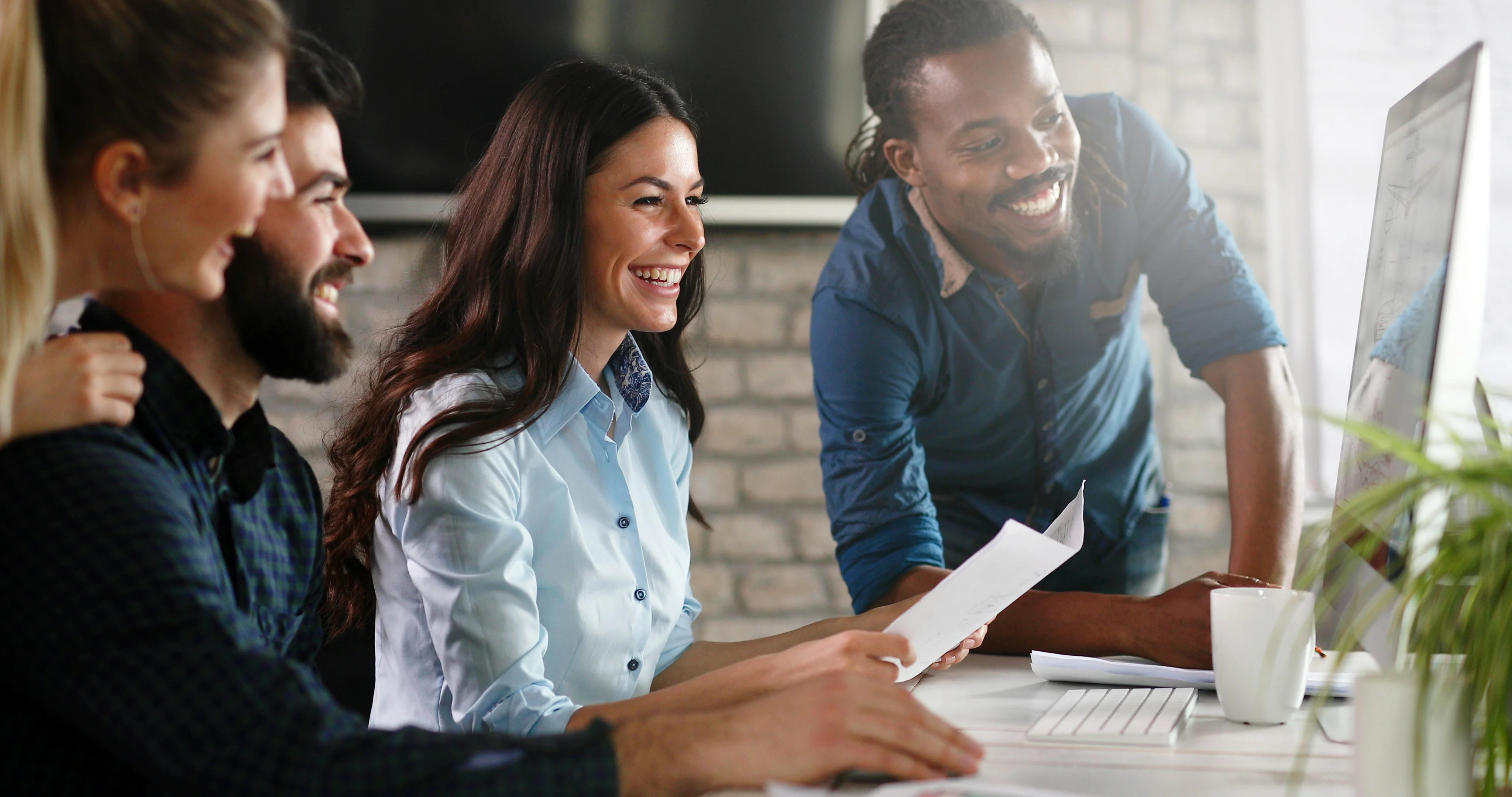 A diverse group of four people smiling and looking at a computer screen in an office setting, with documents and a coffee cup on the table.