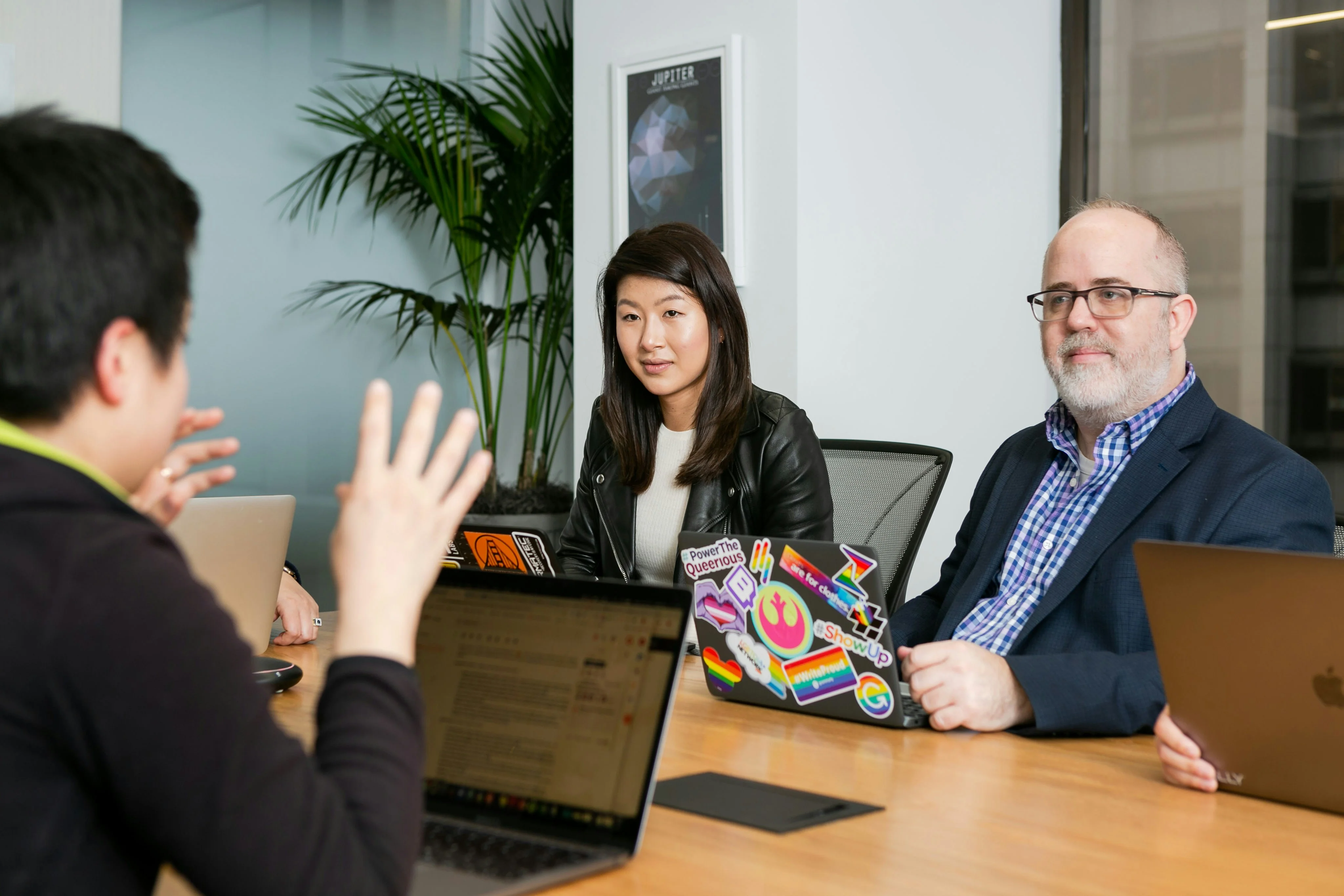 A group of people sit at a conference table with laptops, engaged in discussion. A plant and a poster are visible in the background.