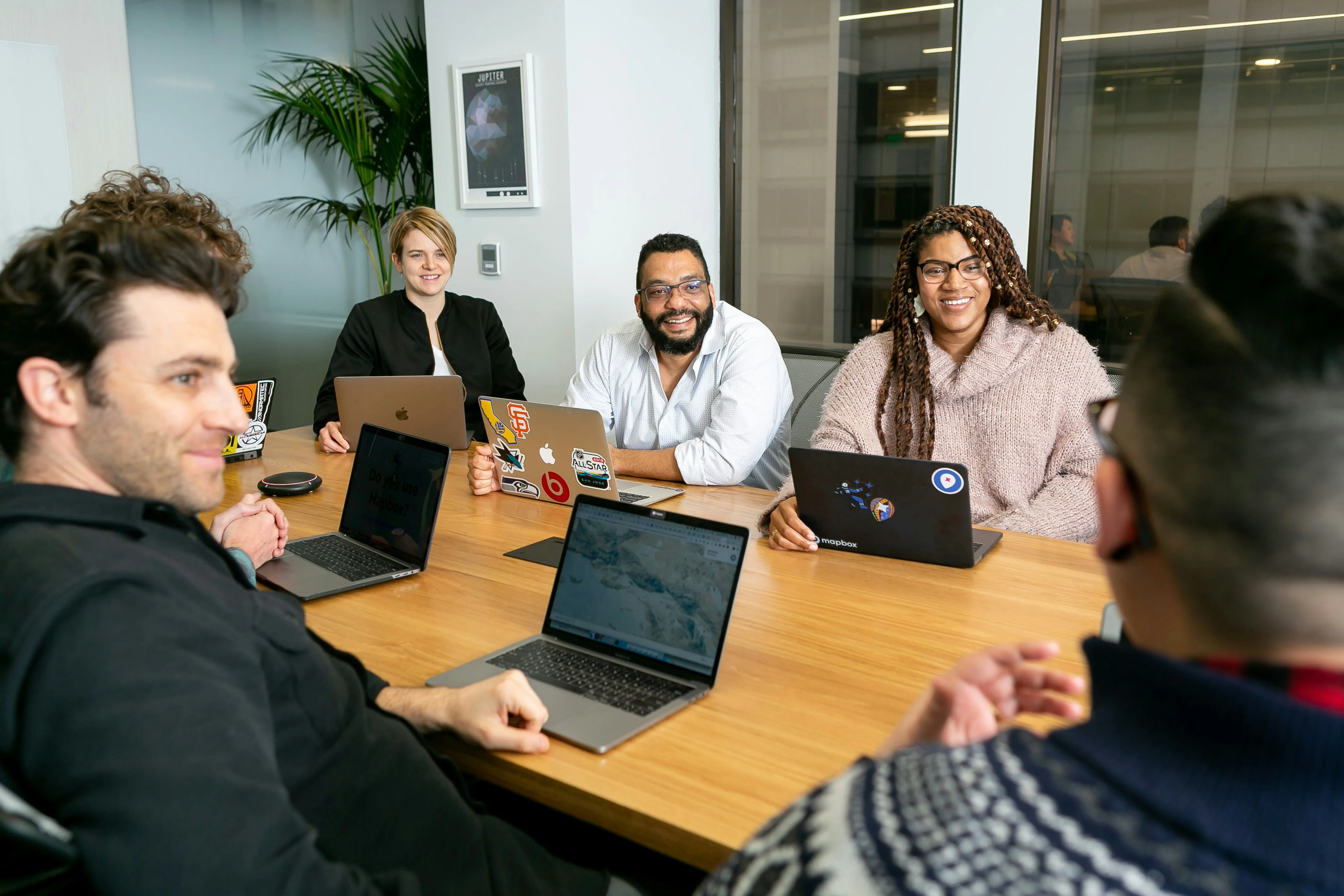A diverse group of people sitting around a conference table with laptops, engaged in a meeting and smiling.