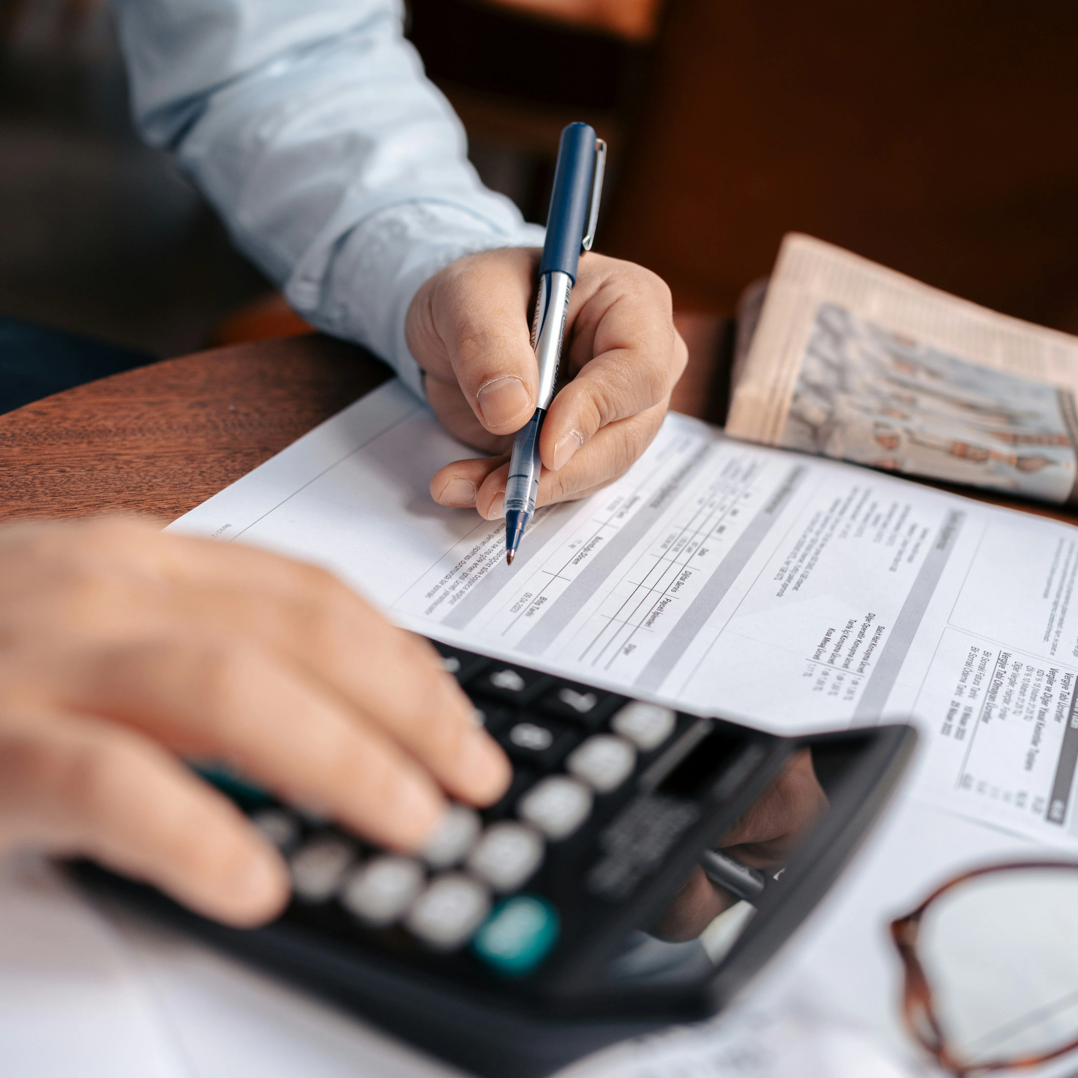 Person using a calculator and writing on documents at a table, with a newspaper and glasses nearby.