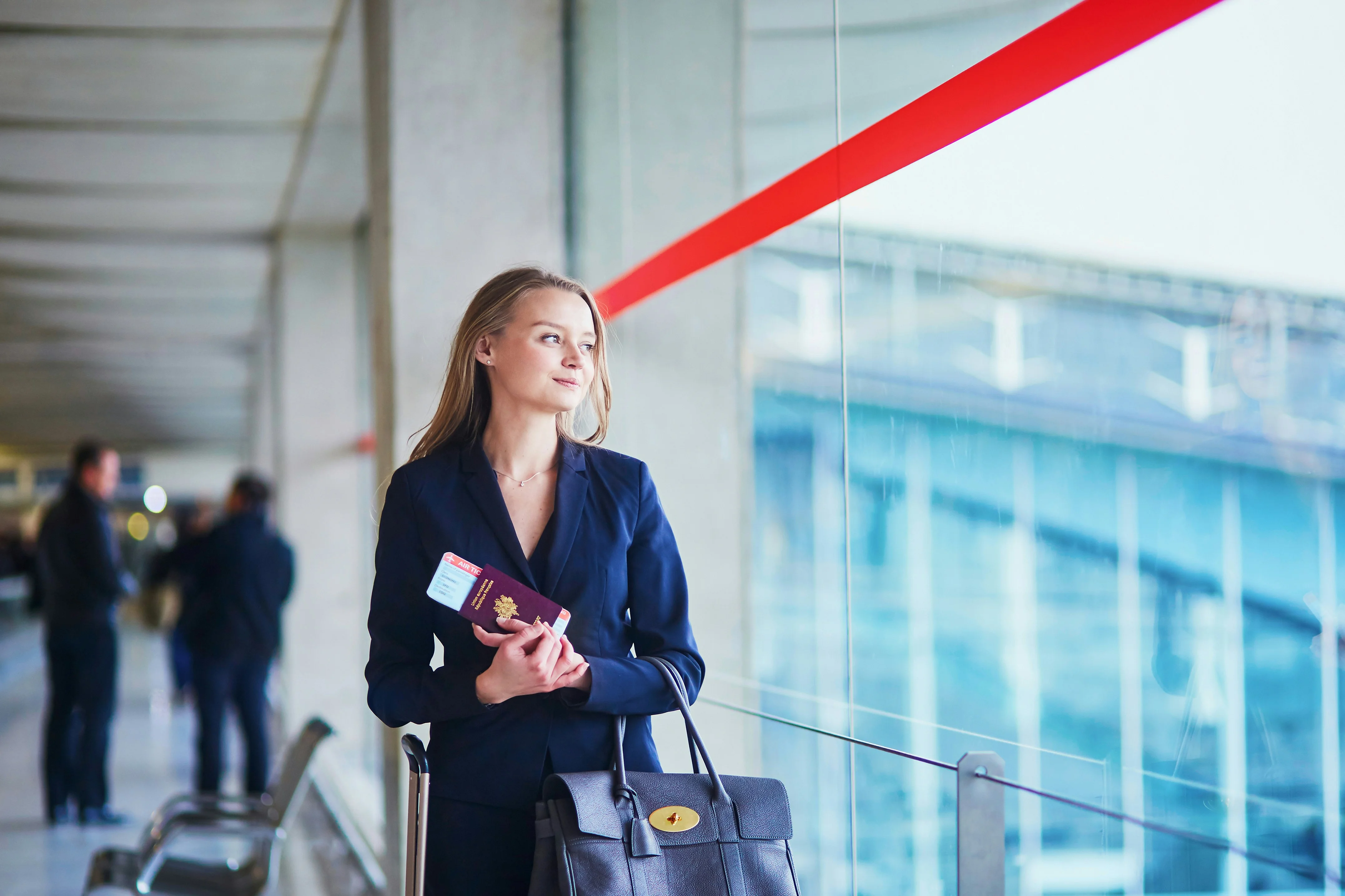 Woman in a blue suit holding a passport and boarding pass, looking out of an airport terminal window with a thoughtful expression.