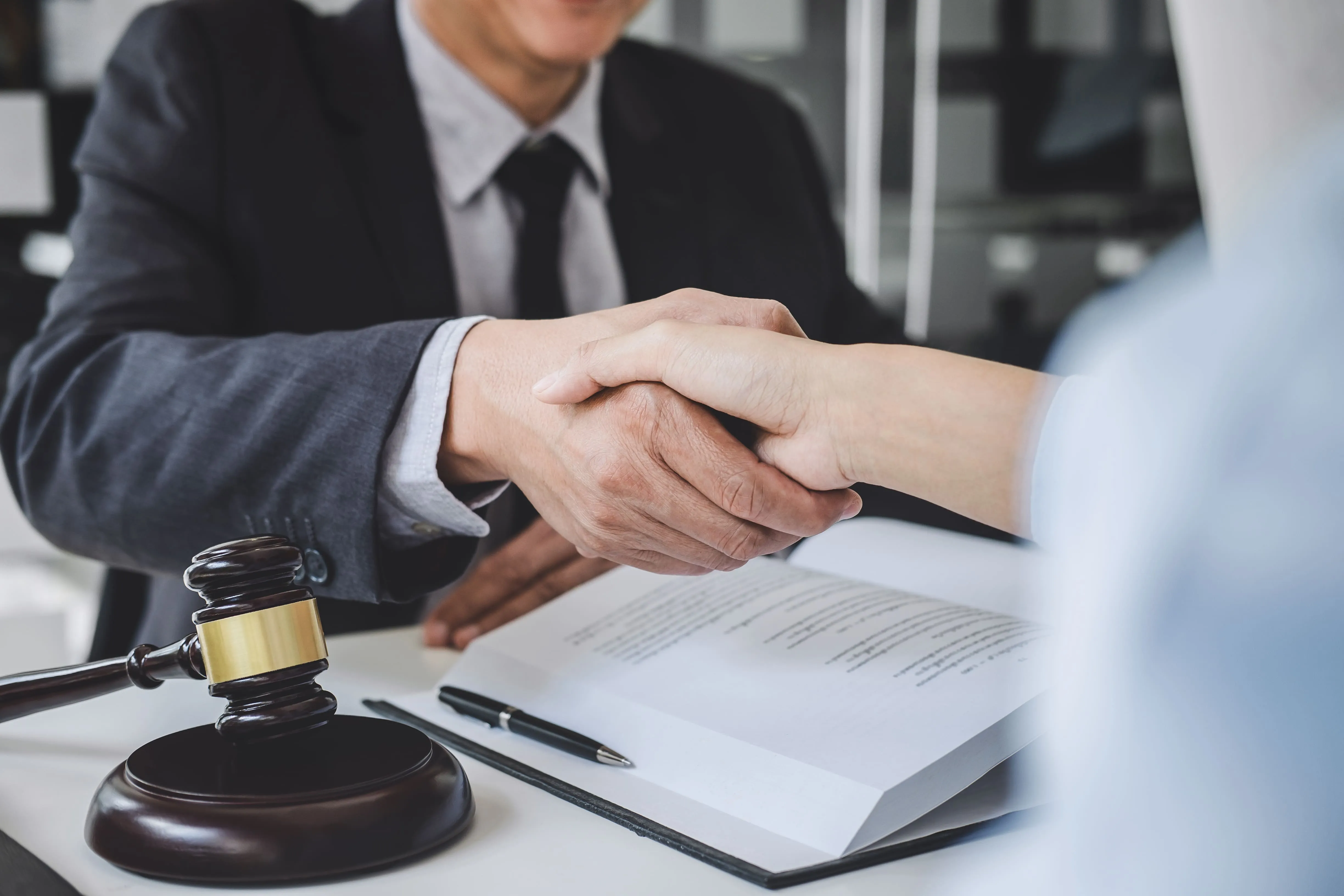 Two people shaking hands over a desk, with a gavel, pen, and open legal document visible.