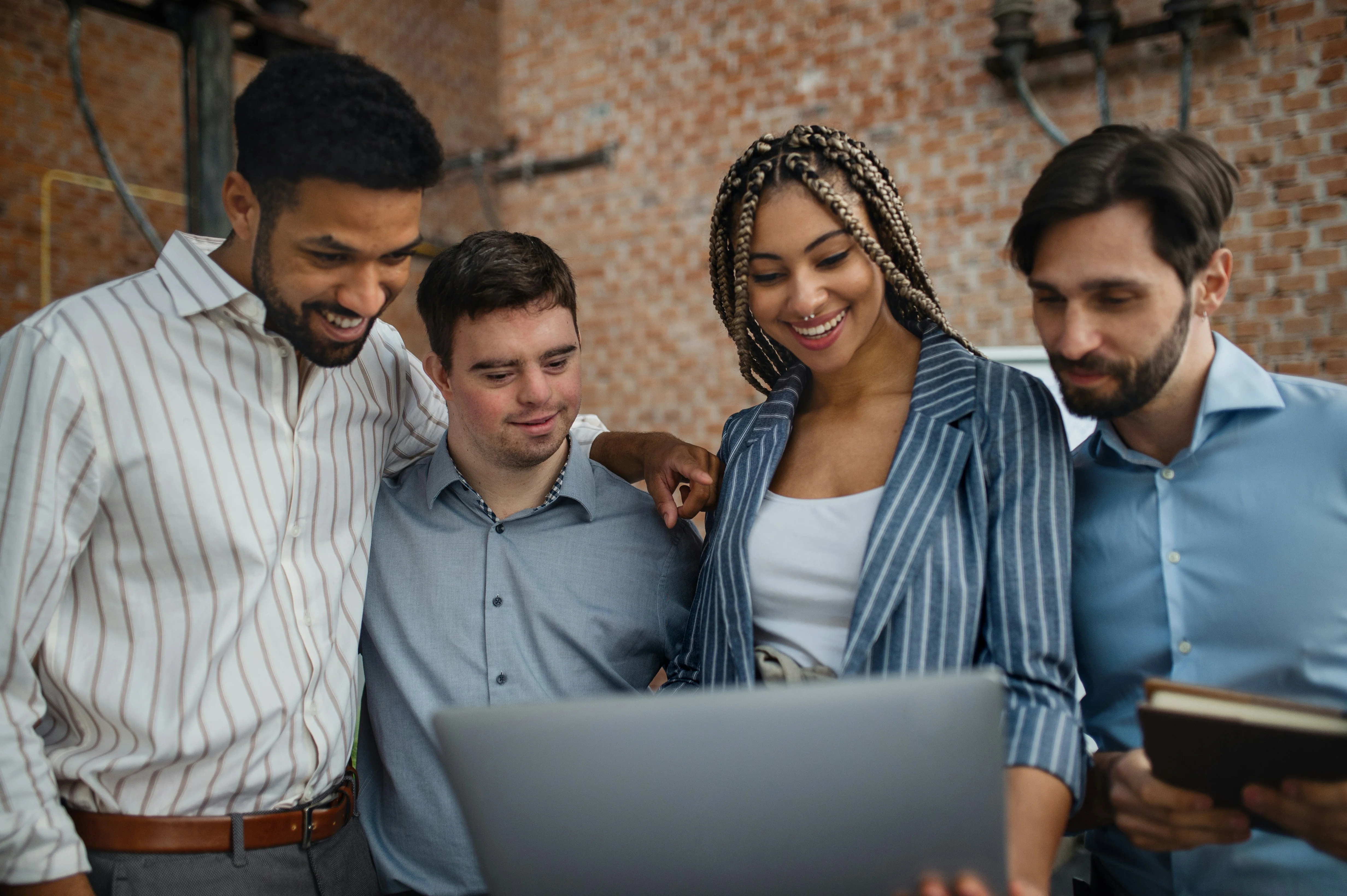 Four people smiling and looking at a laptop together in an office setting, with brick walls in the background.