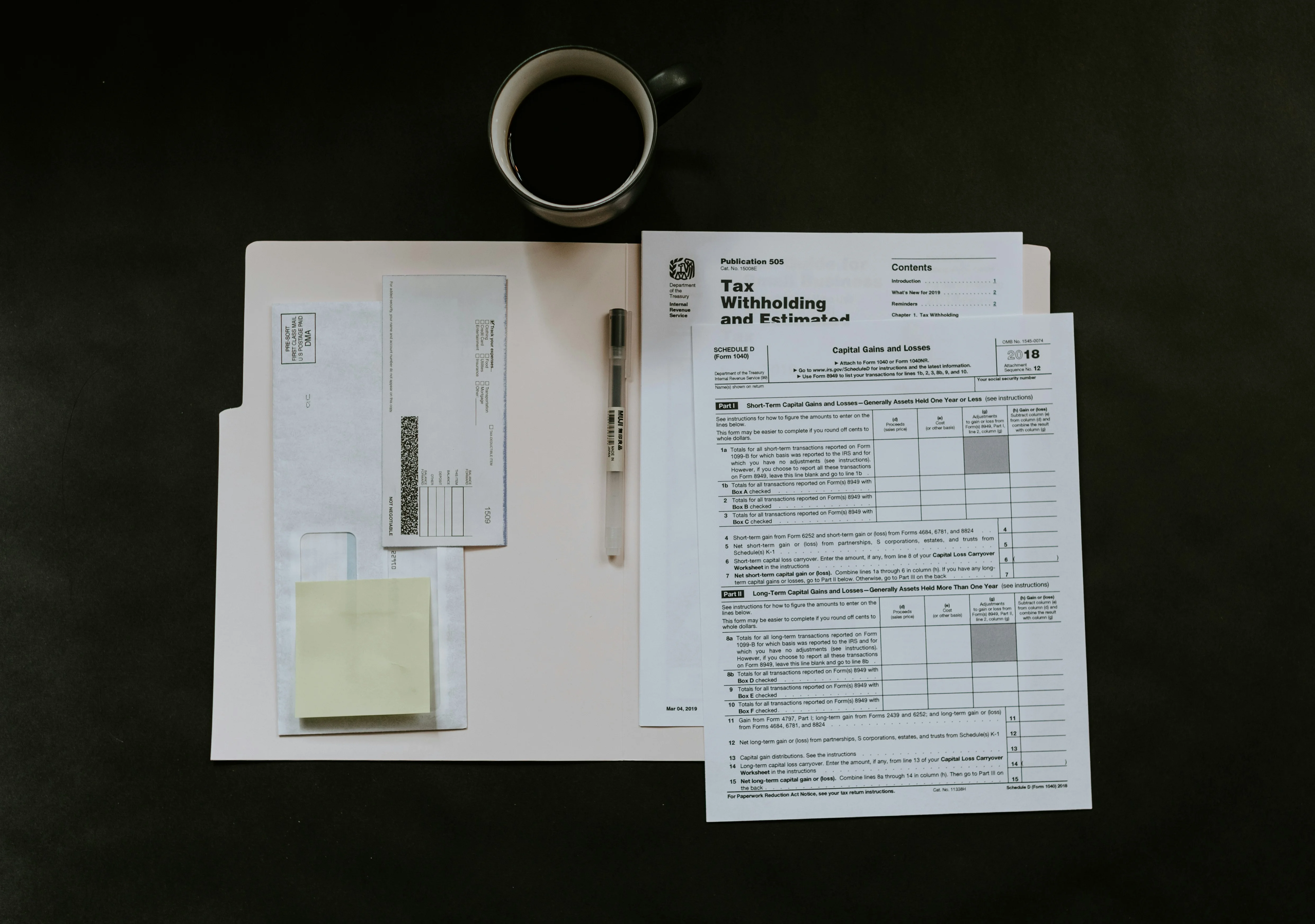 A table with tax documents, a pen, envelopes, sticky notes, and a cup of coffee arranged neatly on a dark surface.