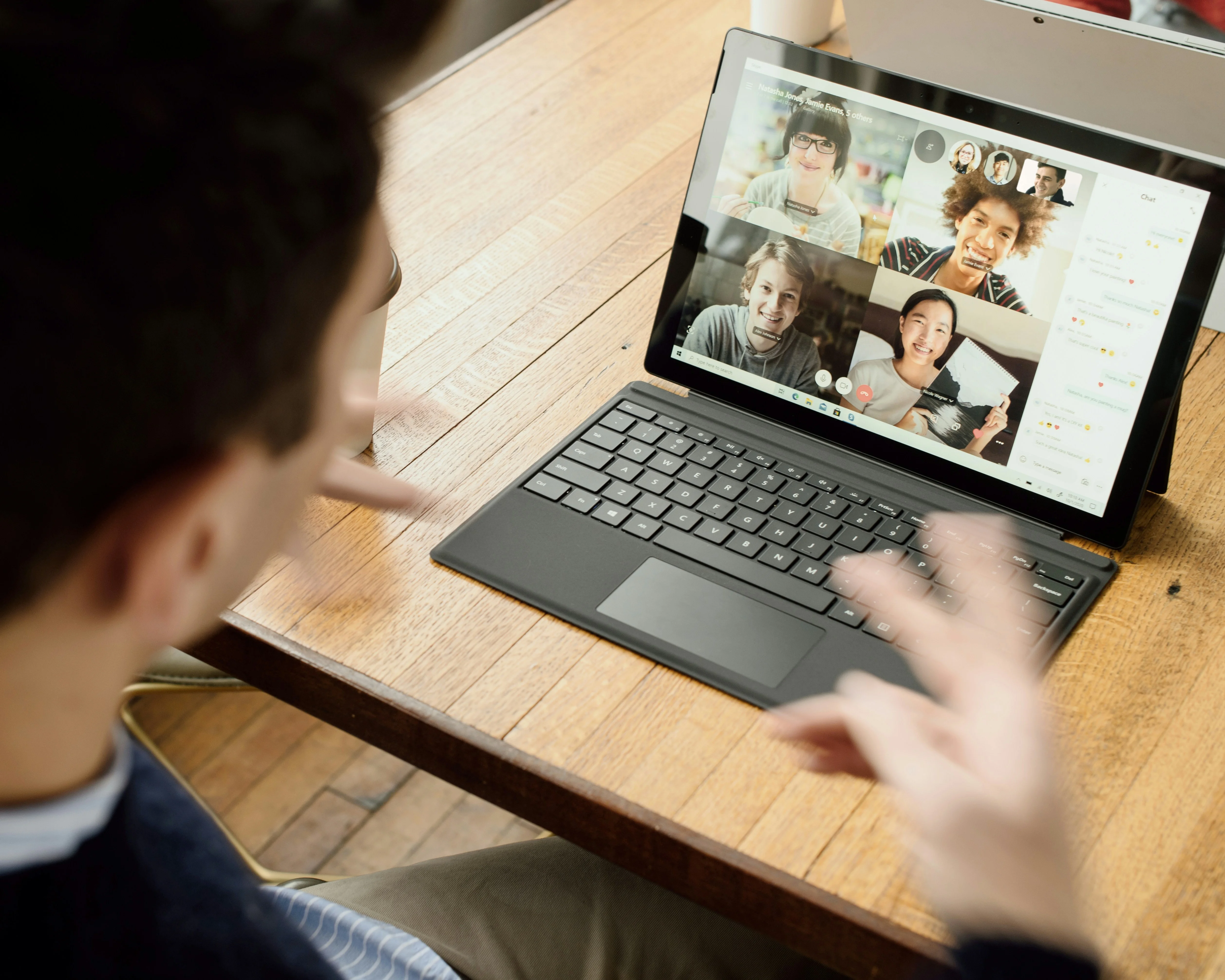 Person sitting at a wooden table, video chatting on a tablet with four people. Hands are gesturing mid-conversation.