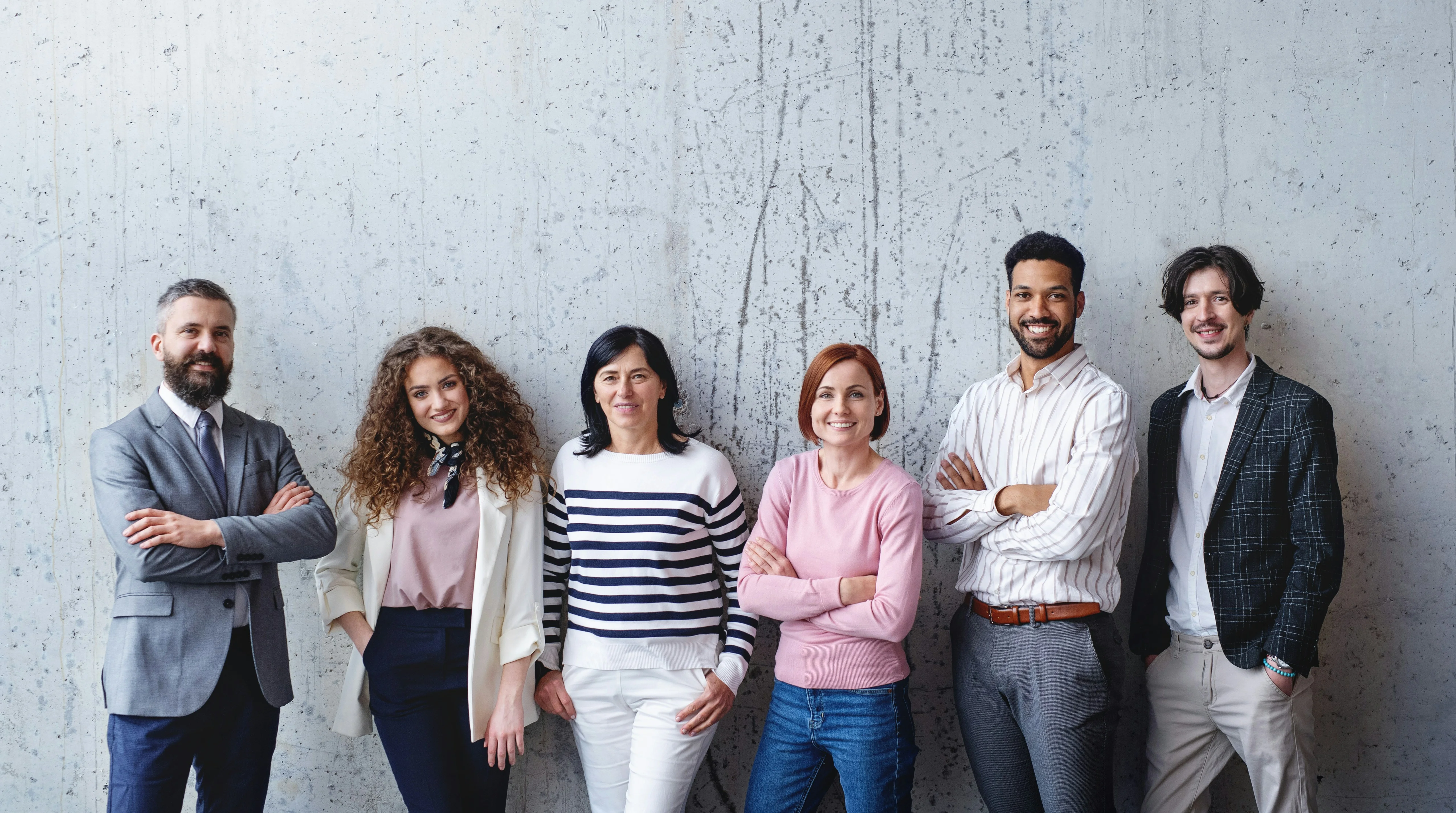 A diverse group of six people standing against a textured wall, smiling with arms crossed, dressed in casual and business attire.