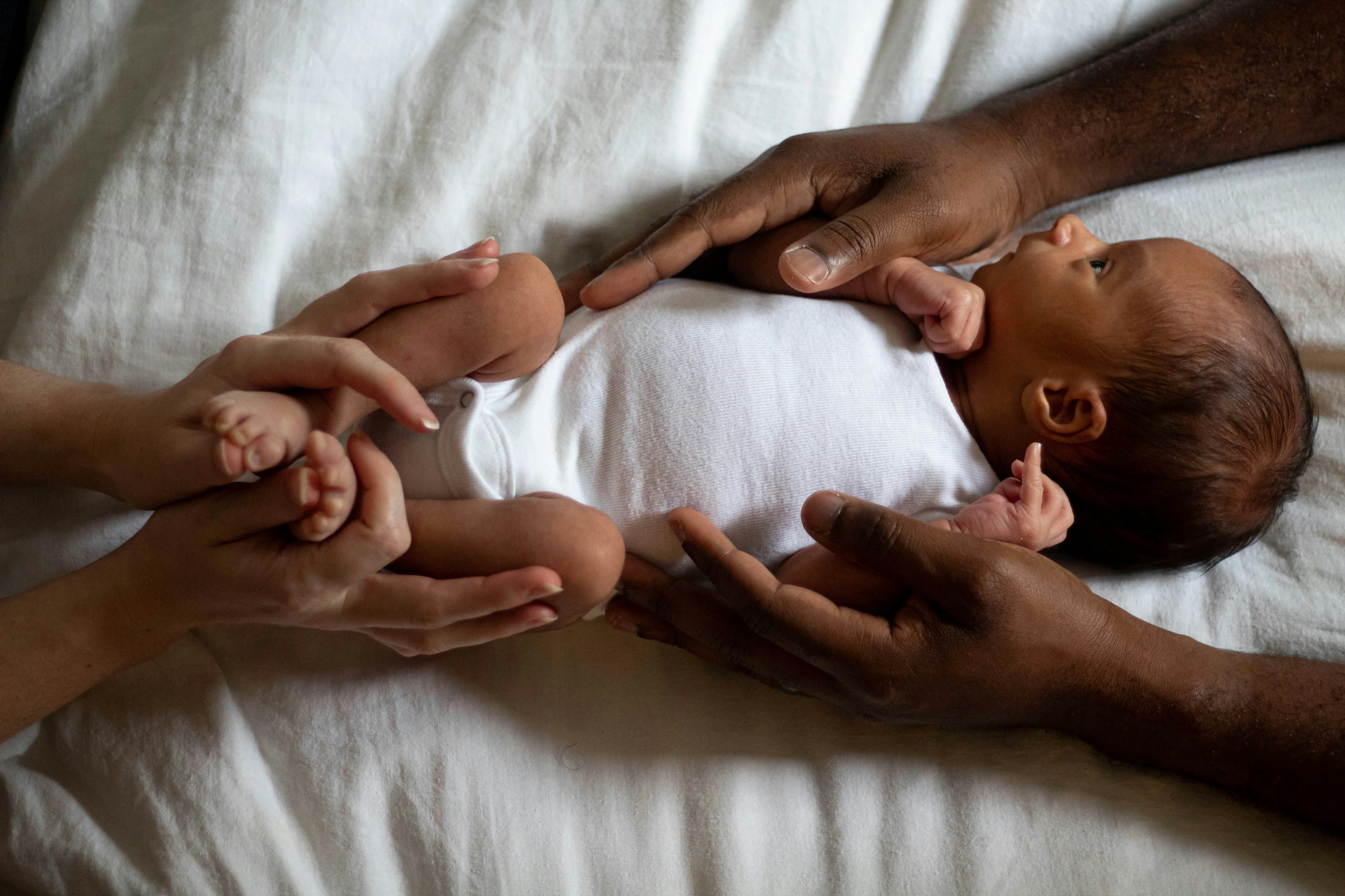 A newborn baby in a white onesie lies on a white surface, surrounded by two pairs of adult hands gently holding and supporting.