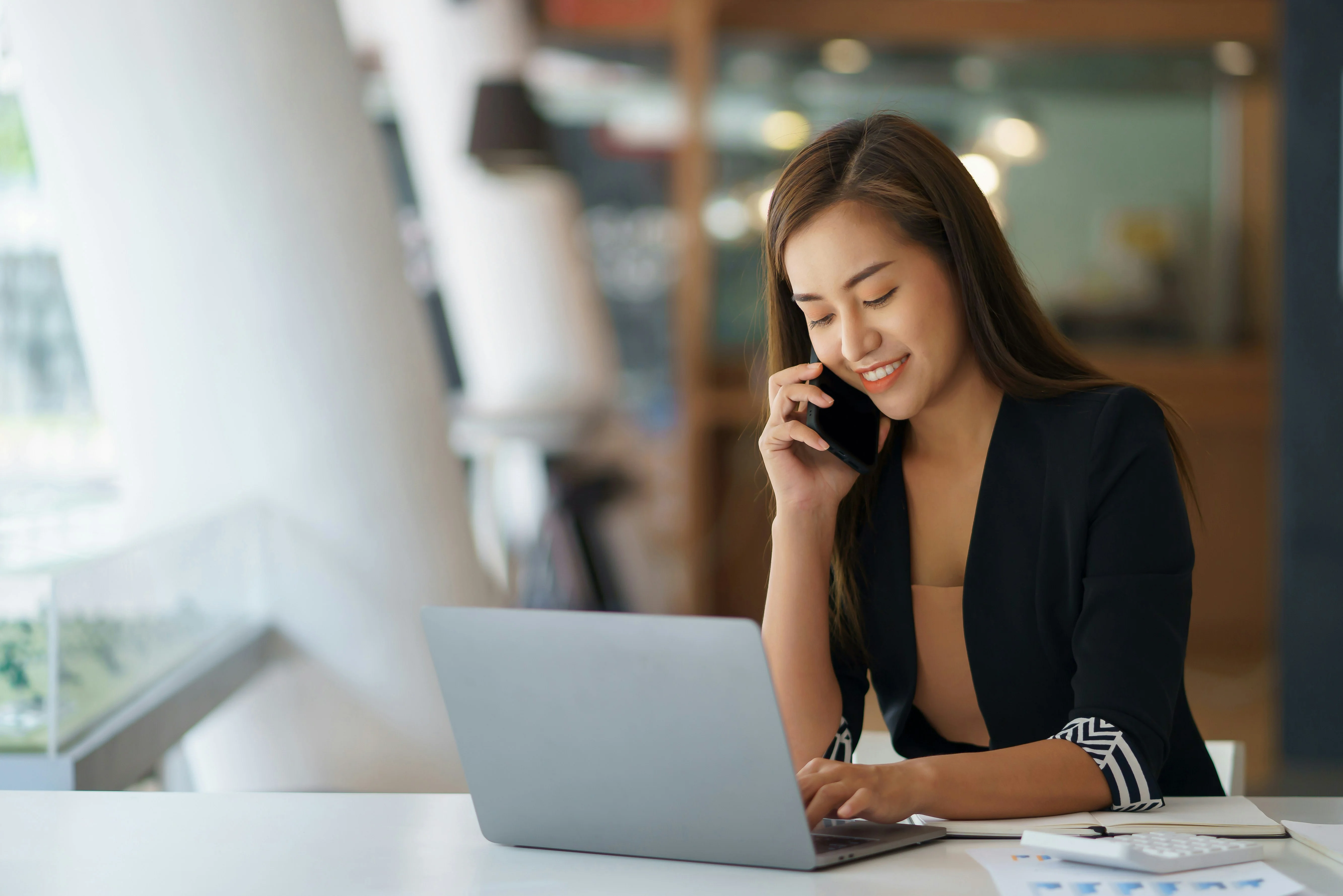 Woman smiling and talking on a phone while working on a laptop at a desk in a modern office setting.