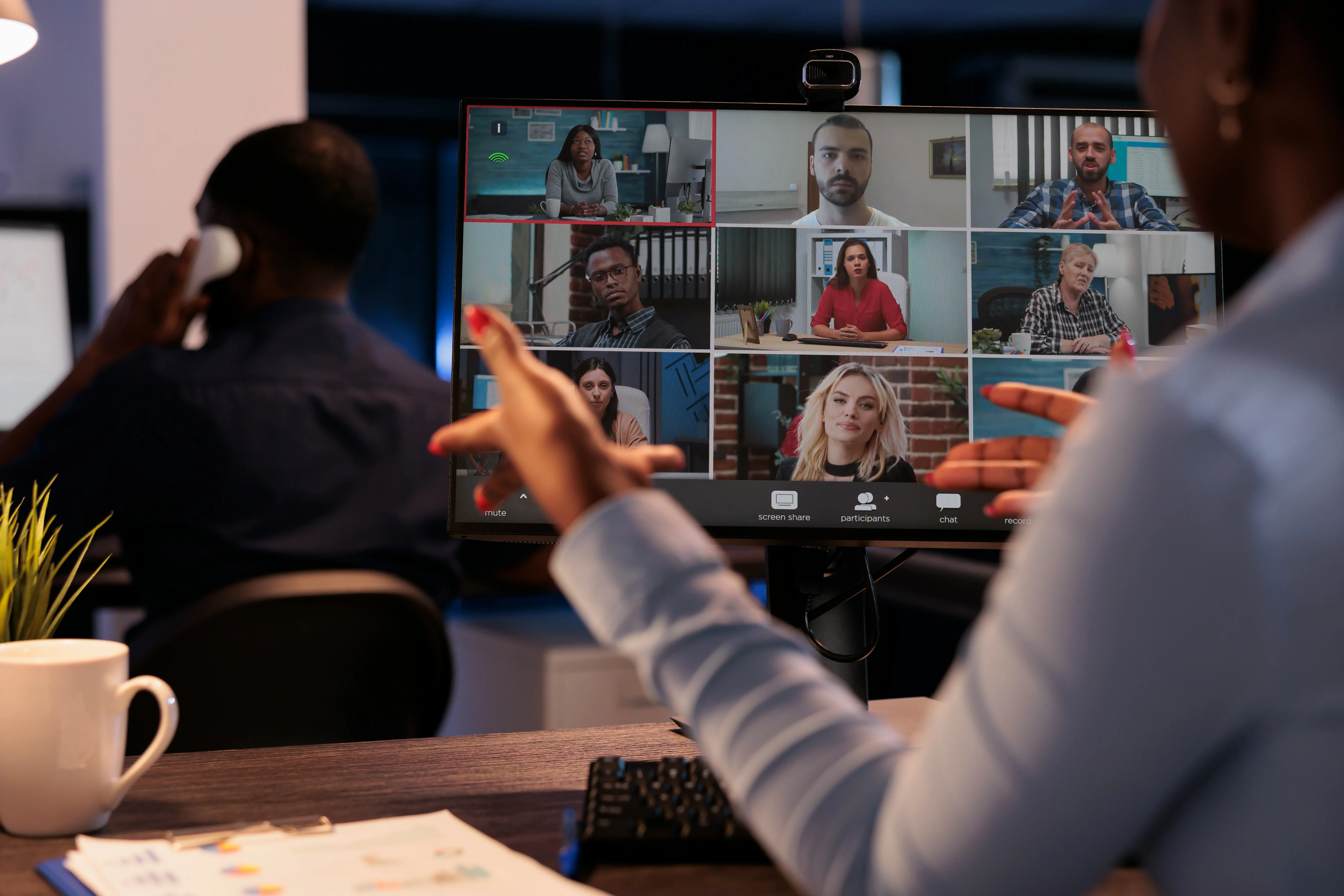 Person gesturing at a monitor showing a nine-person video conference grid, with a desk, keyboard, coffee mug, and papers in the foreground.