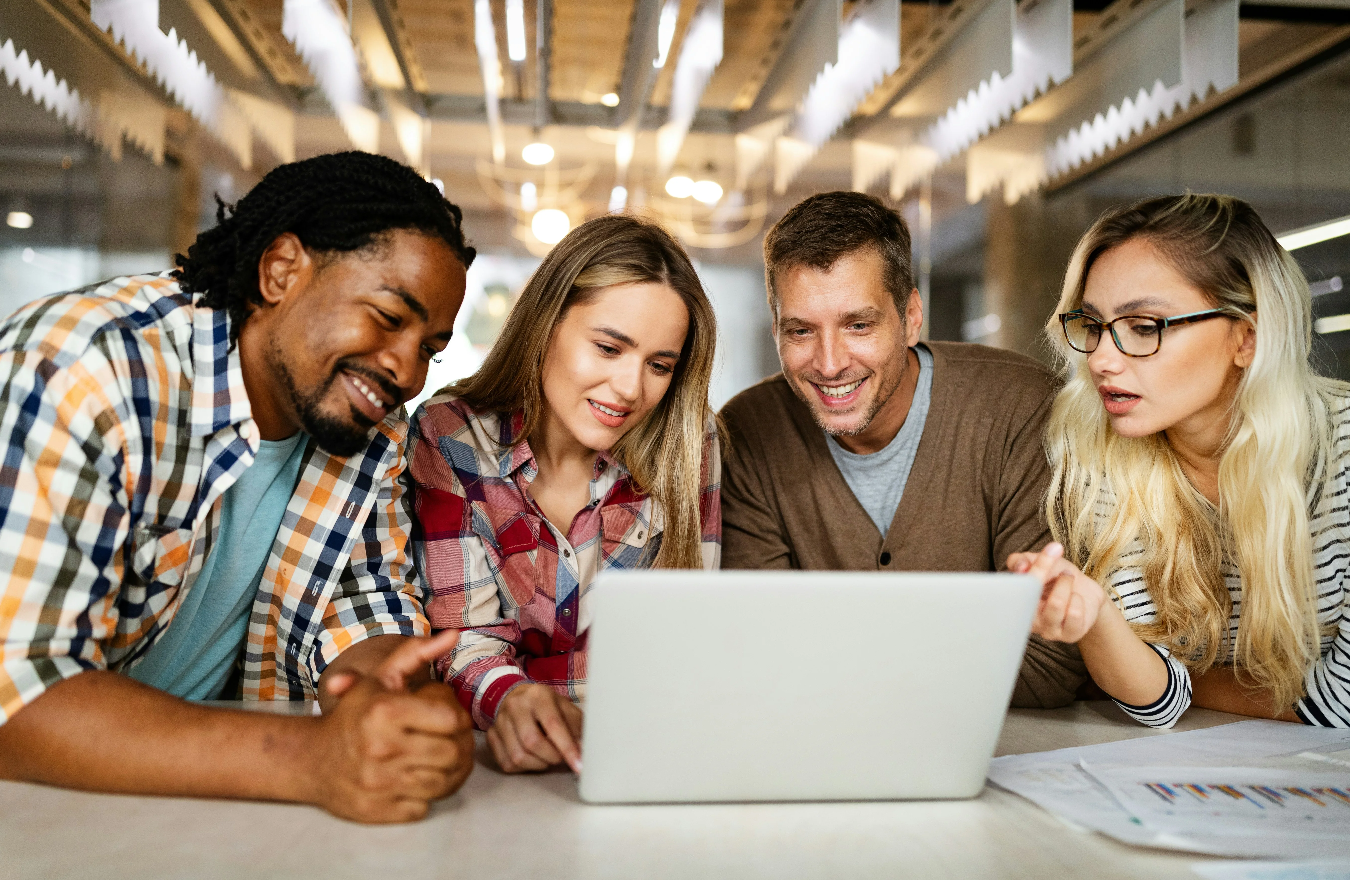 Four people smiling and looking at a laptop screen together in a modern office setting.