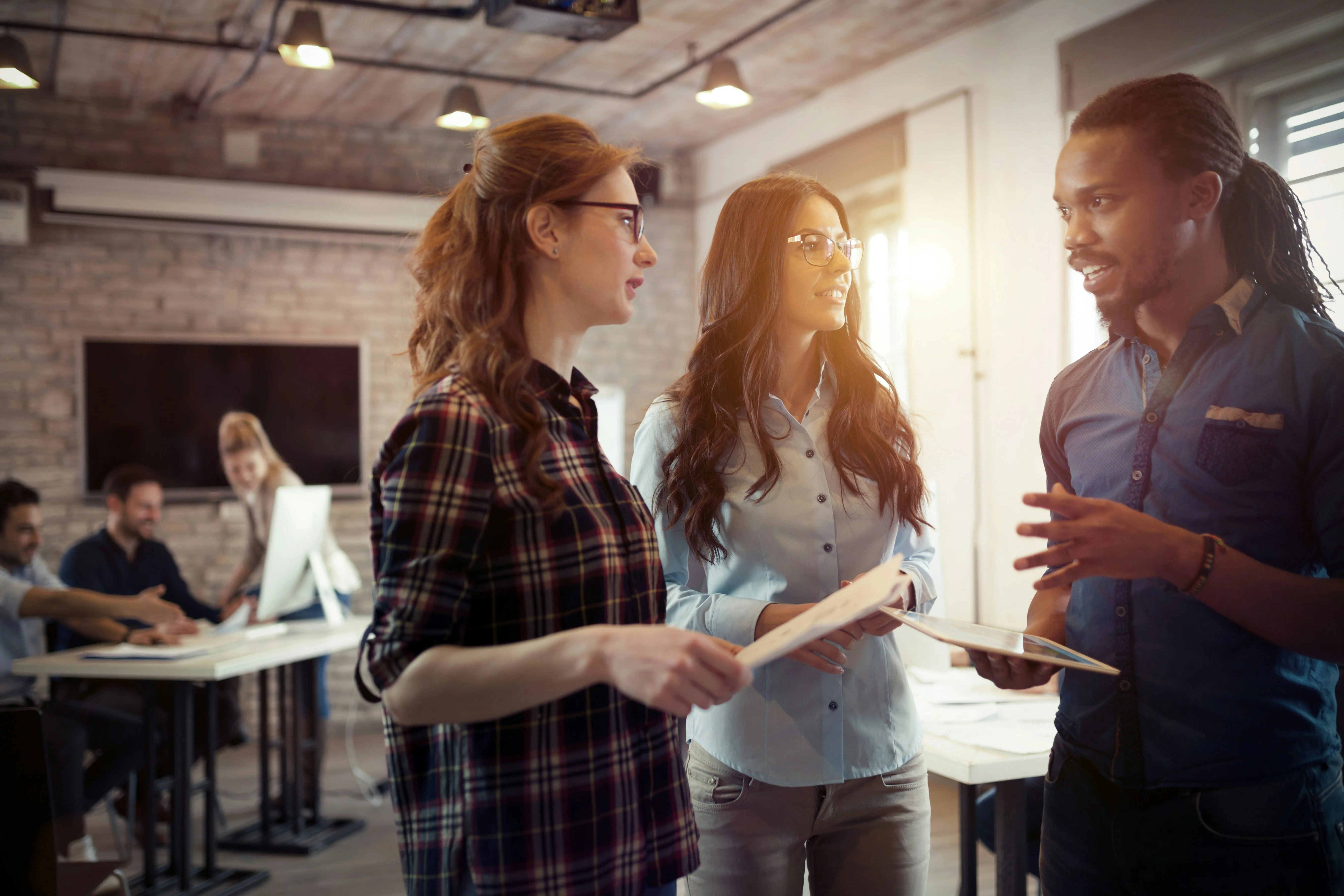 Three colleagues discussing work in a modern office with exposed brick walls and natural lighting, while others work in the background.