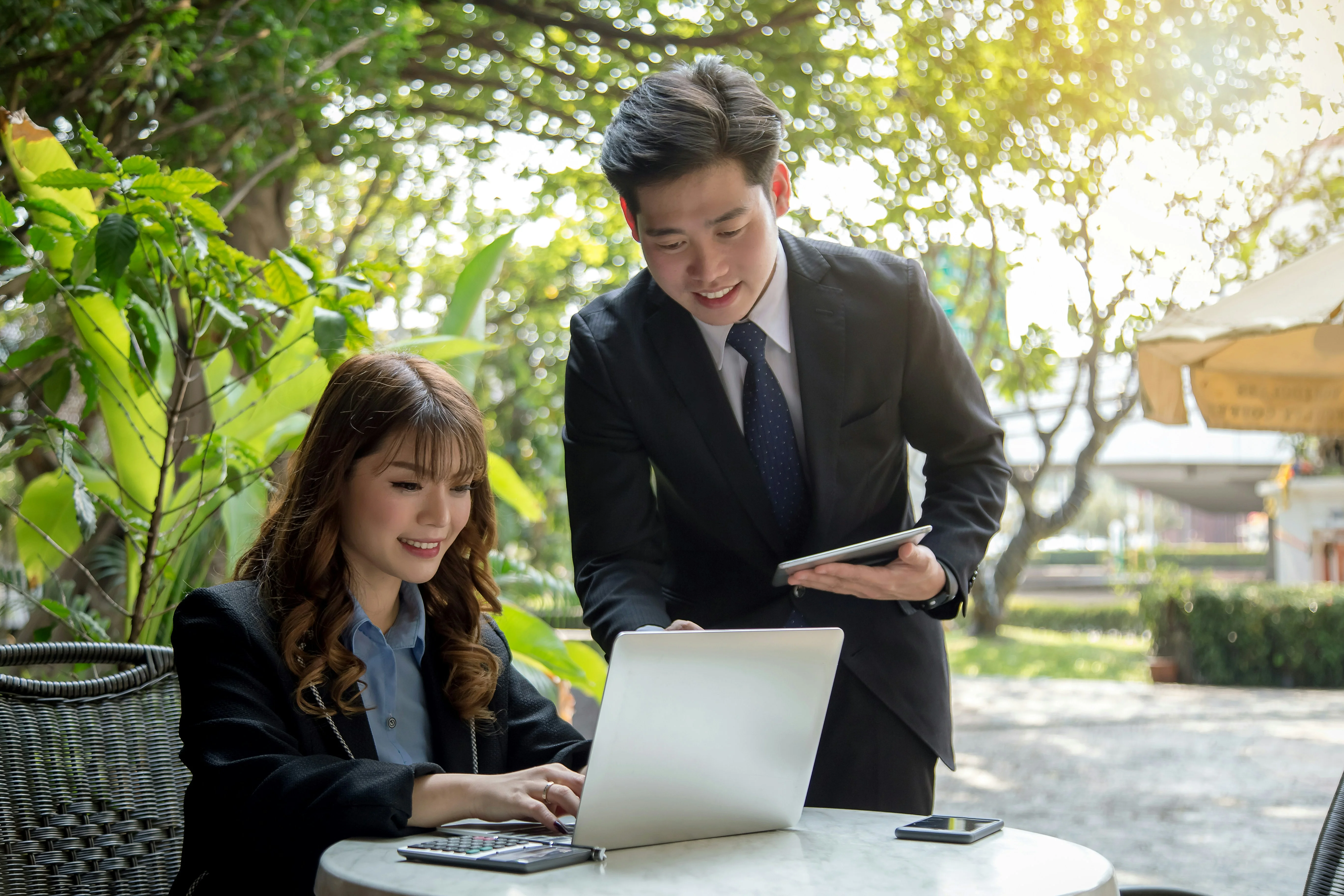 Two business professionals in suits collaborate outdoors, one using a laptop and the other holding a tablet, in a sunlit, leafy setting.