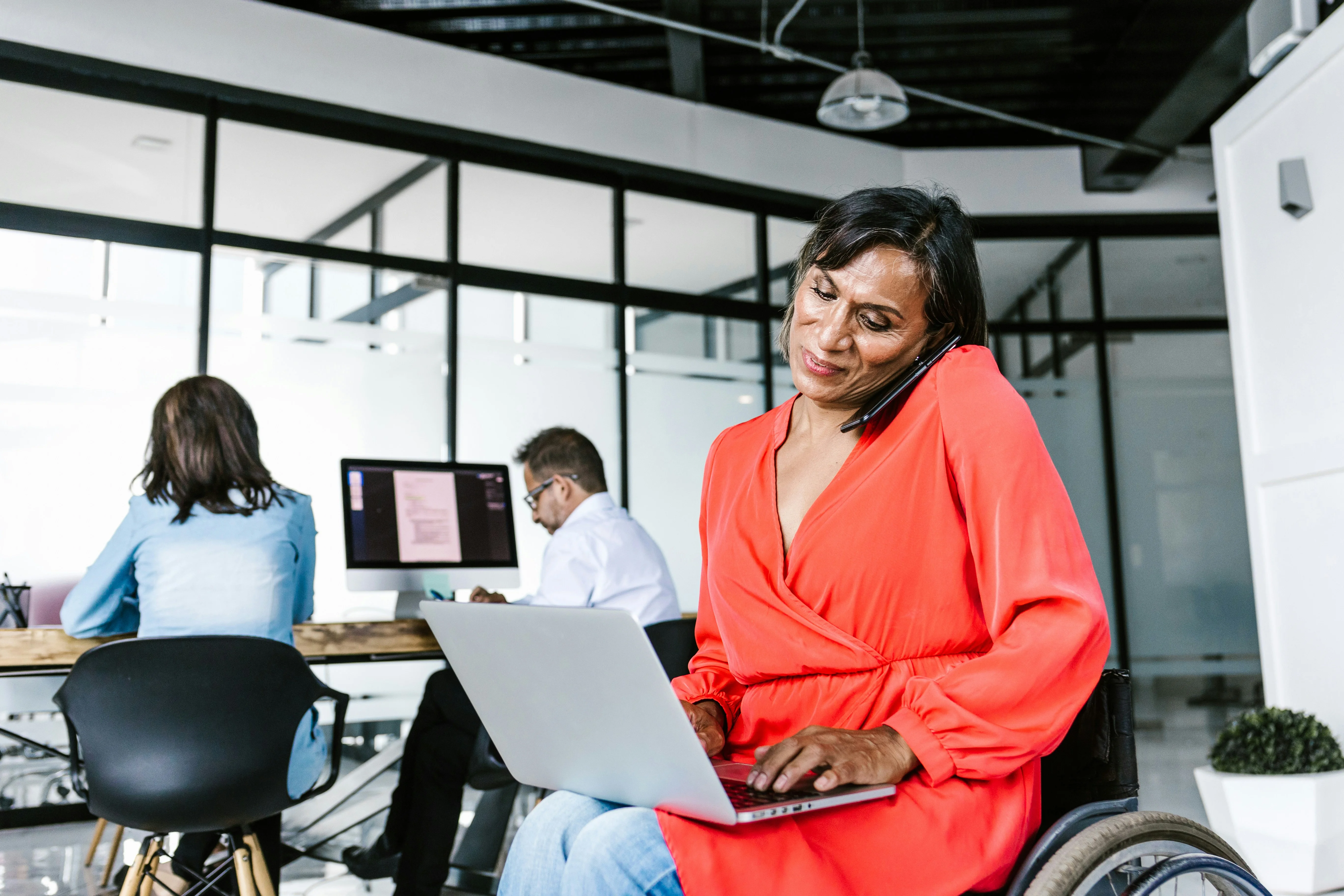 Woman in a wheelchair in a modern office, talking on phone while using a laptop, coworkers working at desks behind her.