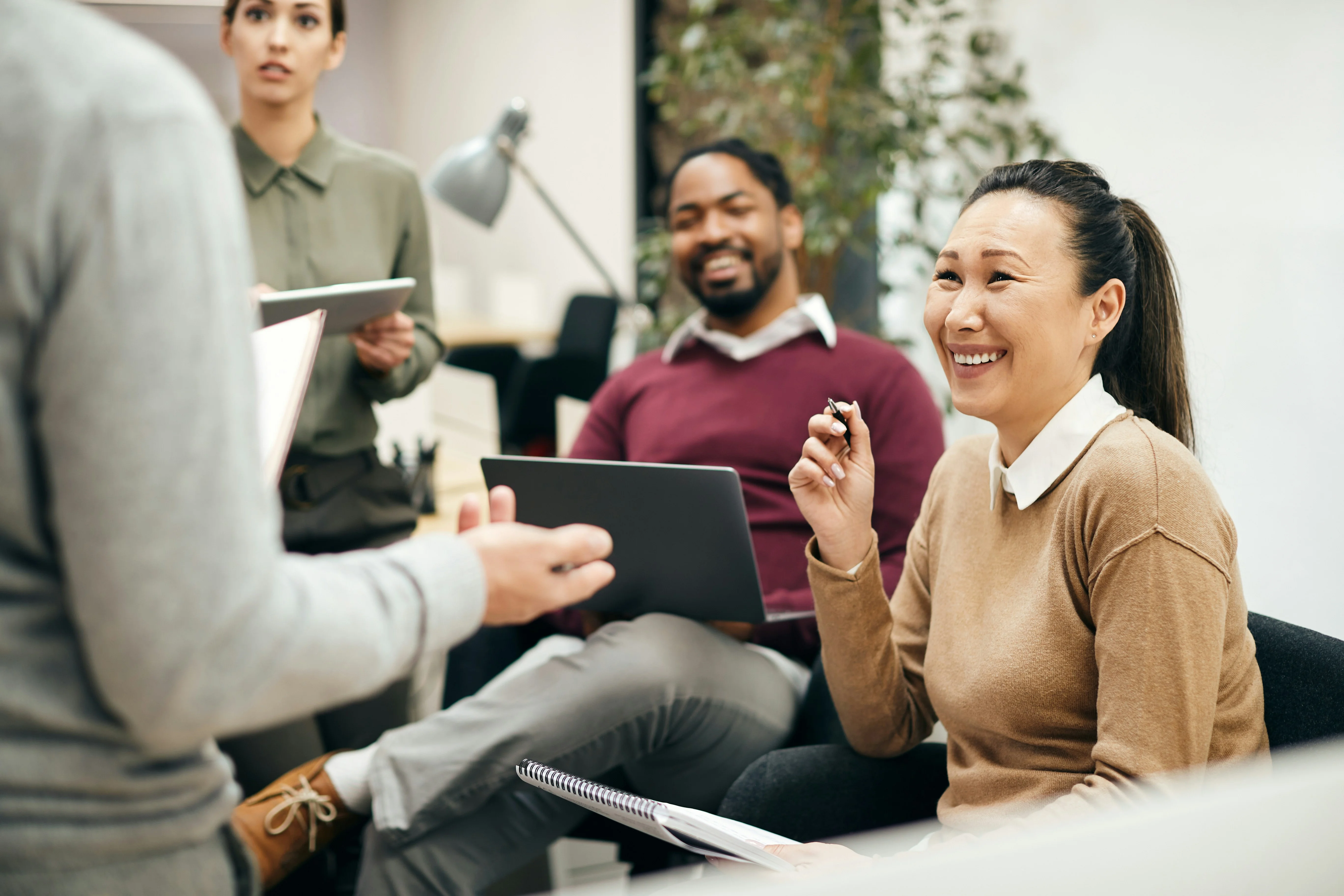 A group of people having a meeting in an office. A woman smiles while holding a pen, and a man with a laptop sits nearby, listening attentively.