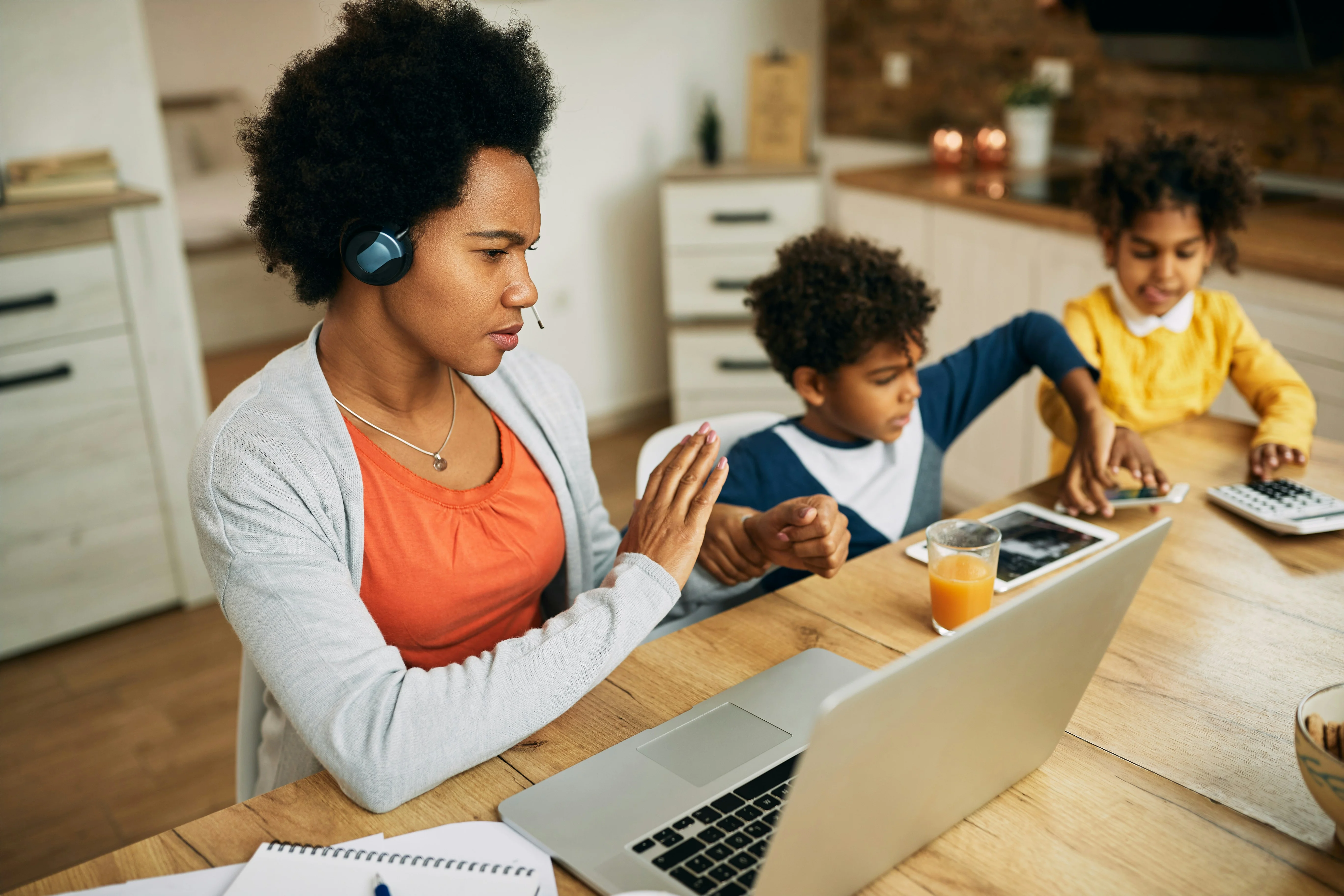 Woman working on a laptop at a kitchen table, wearing a headset. Two children beside her using a tablet and a calculator.