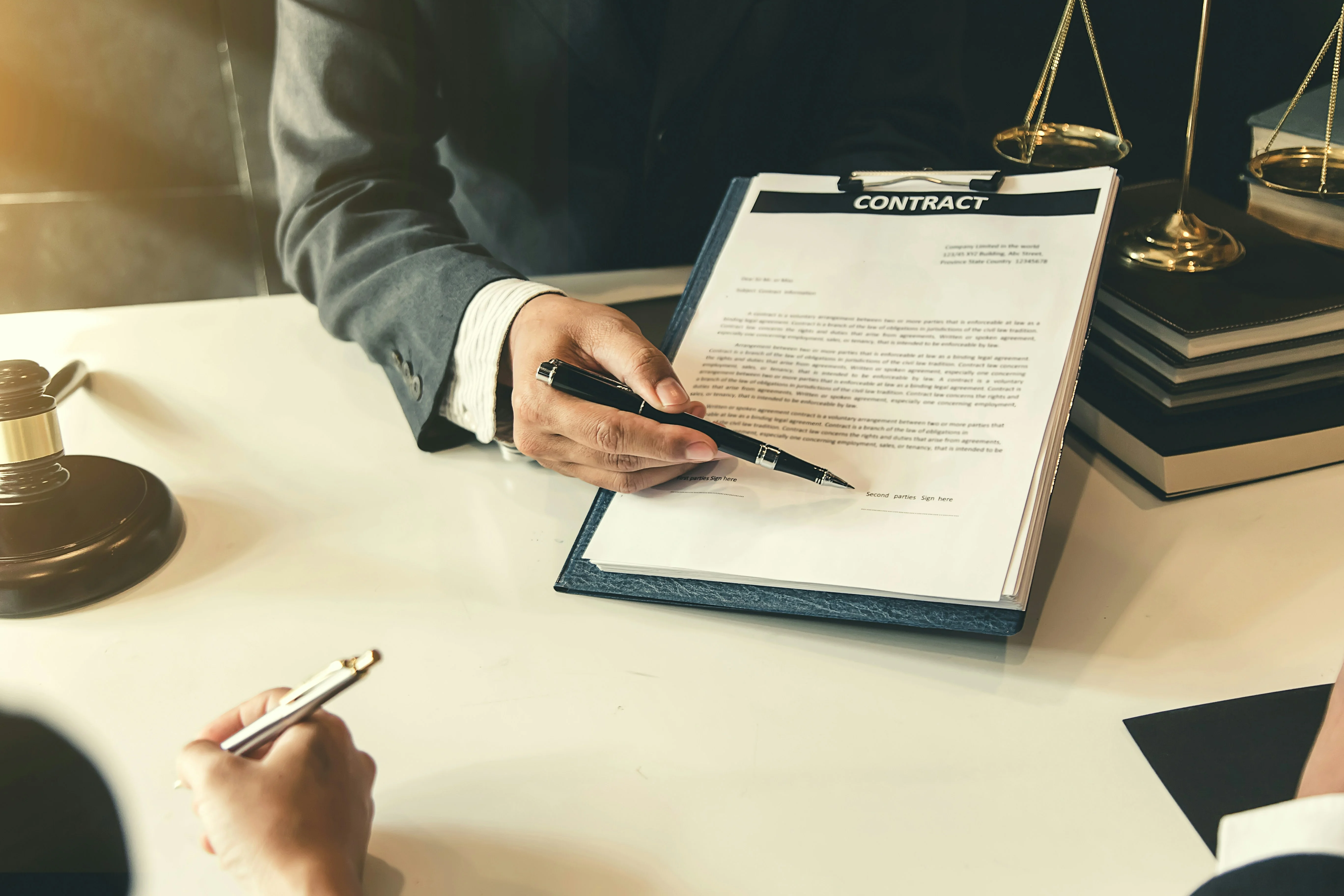 Two people at a desk with legal documents, a pen, and a justice scale, discussing a contract.