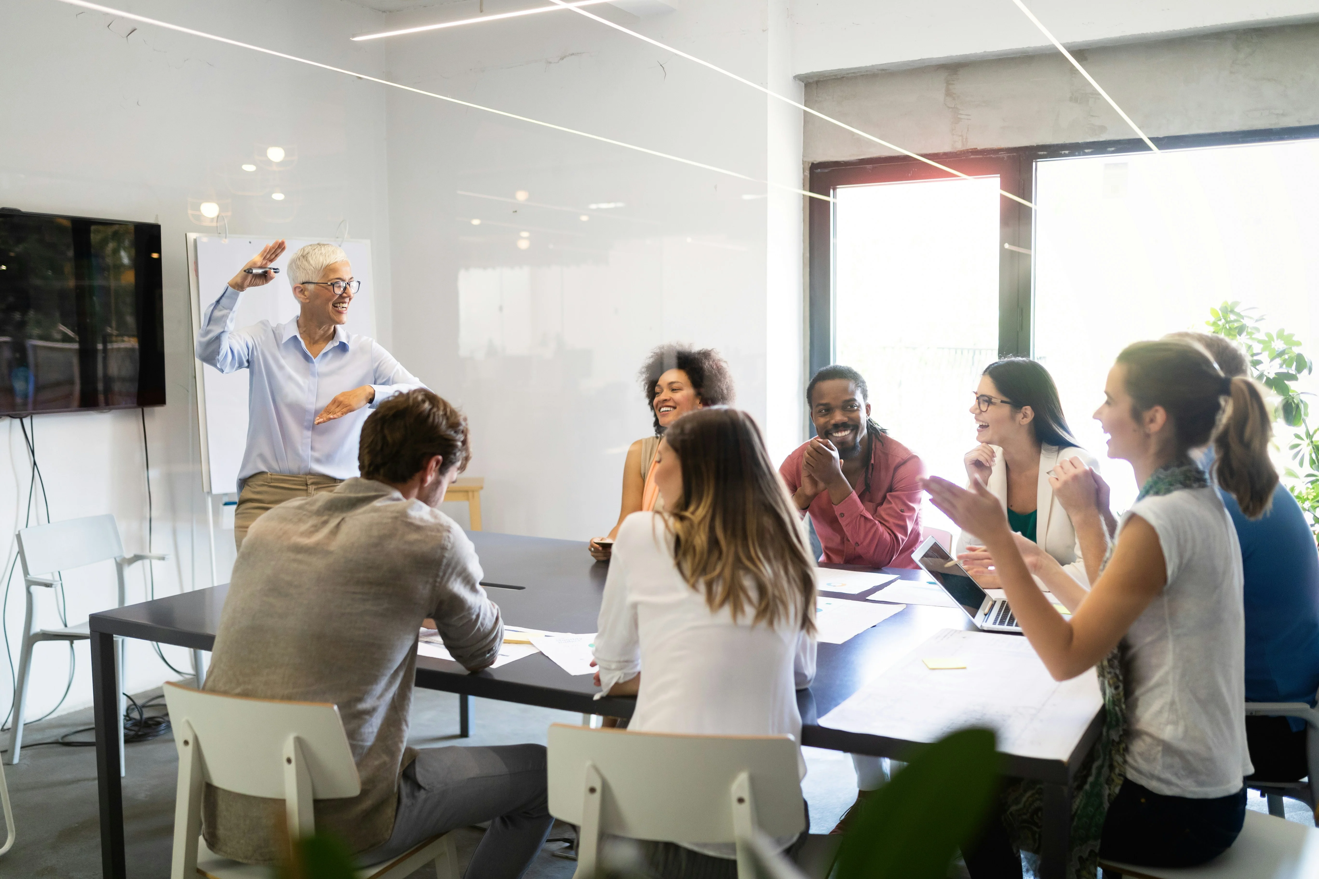 A diverse group of people engaged in a lively meeting around a table in a modern office setting, with one person standing and gesturing.