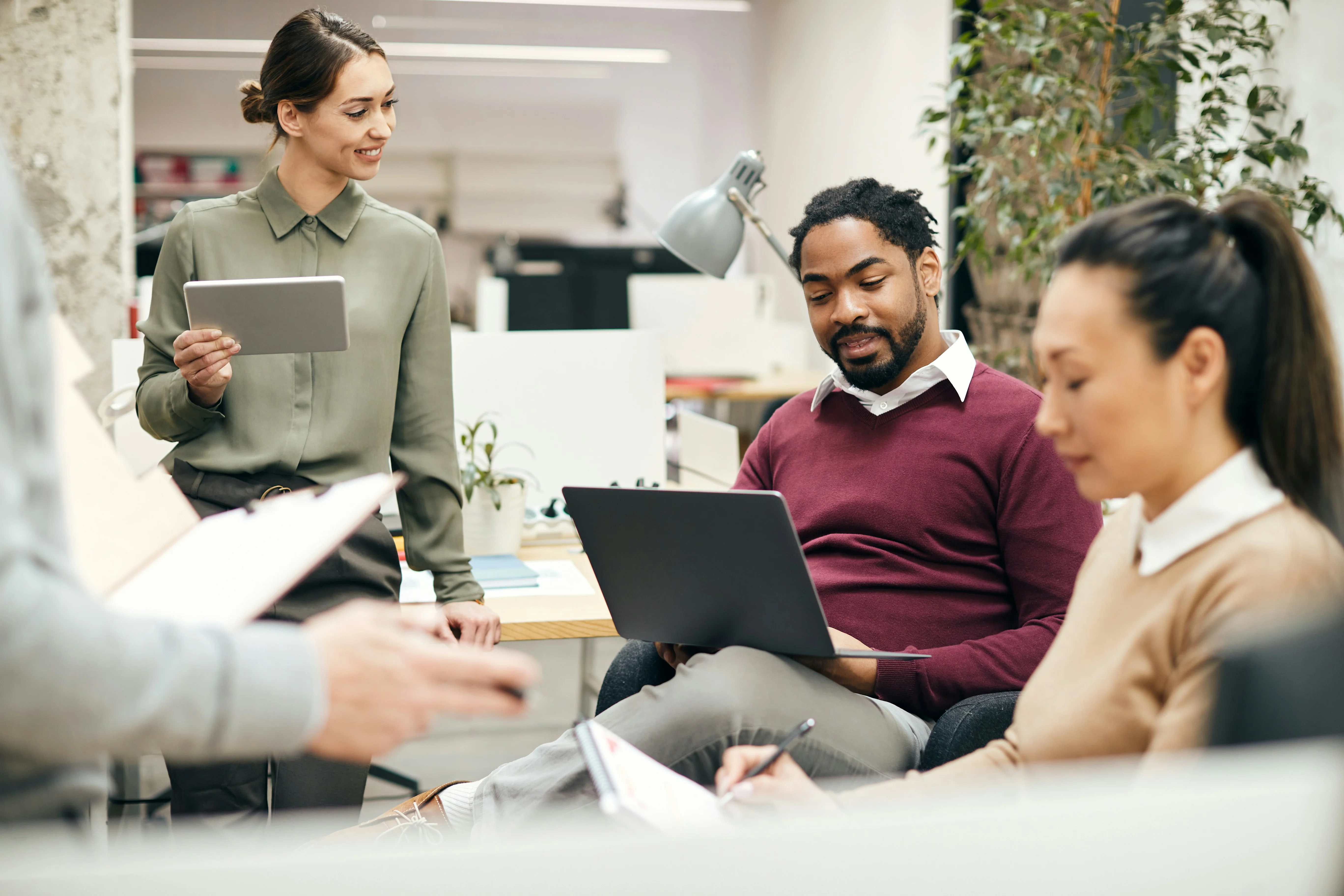 Three colleagues in an office setting, engaged in discussion. One holds a tablet, another a laptop, and the third takes notes.