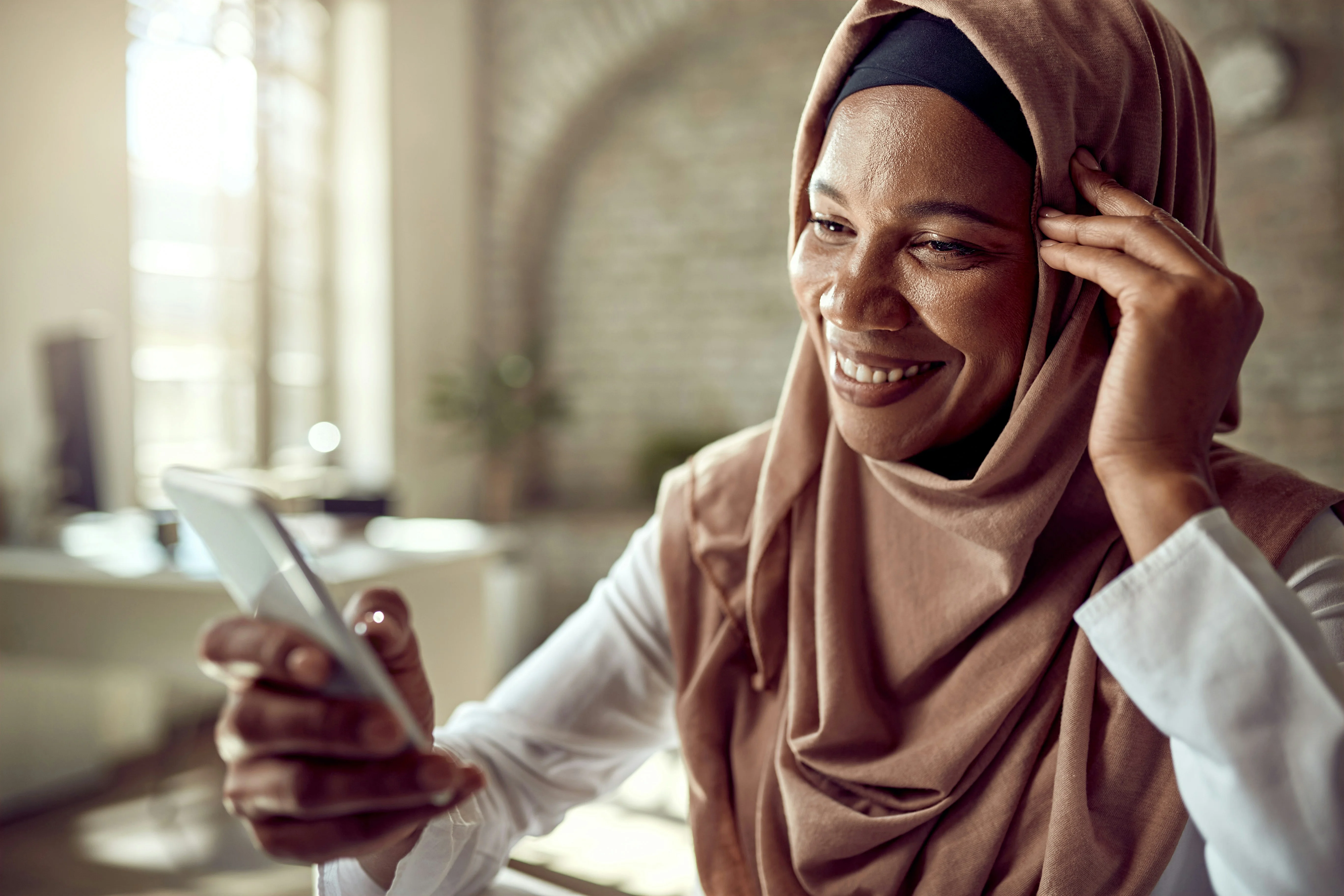 Smiling woman in a hijab holds a smartphone, sitting indoors with soft natural light and blurred background.