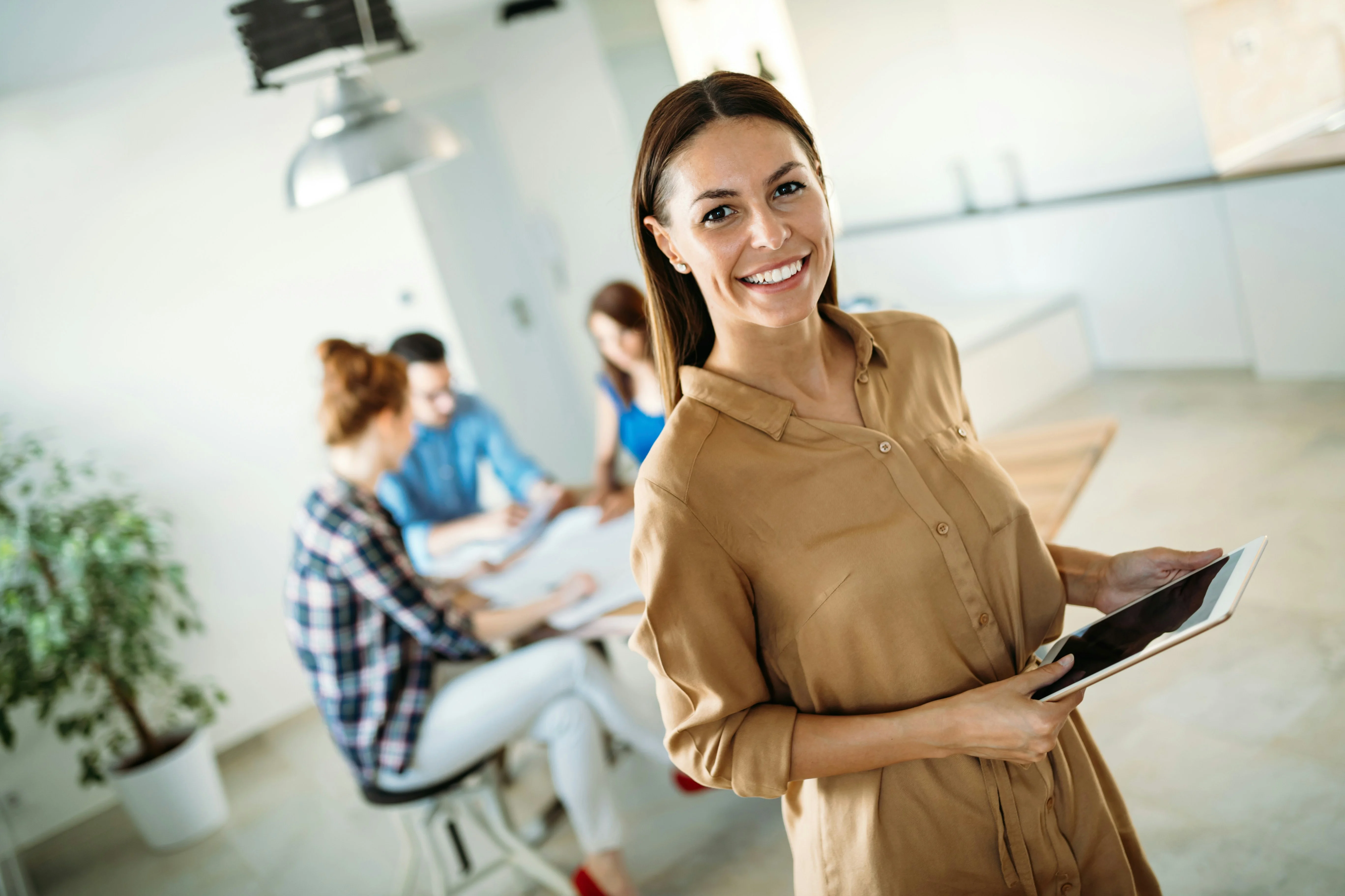 Smiling woman holding a tablet in a bright office, with three colleagues working at a table in the background.