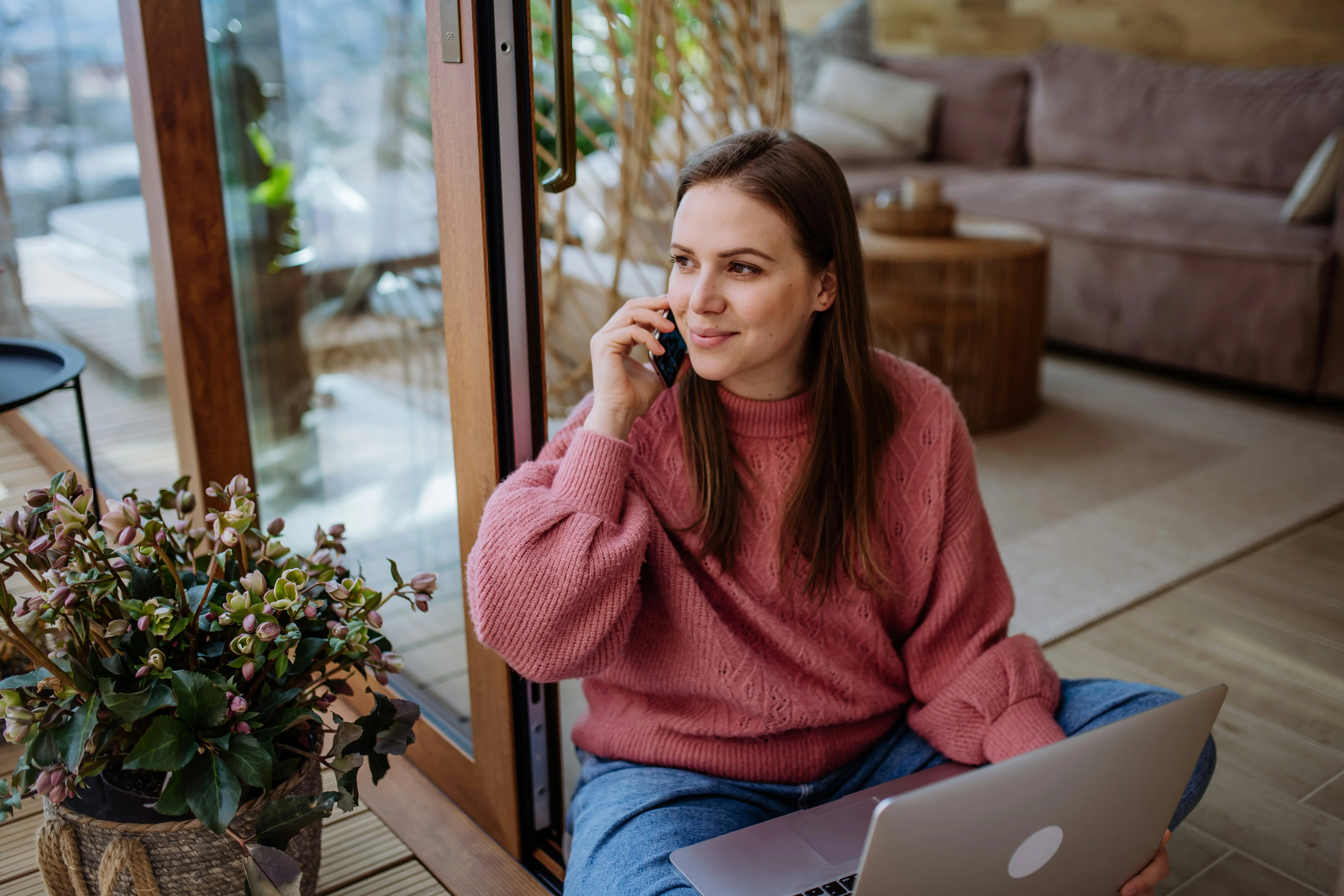 Woman in a pink sweater sits on the floor by a window, talking on the phone and using a laptop, with a potted plant nearby.