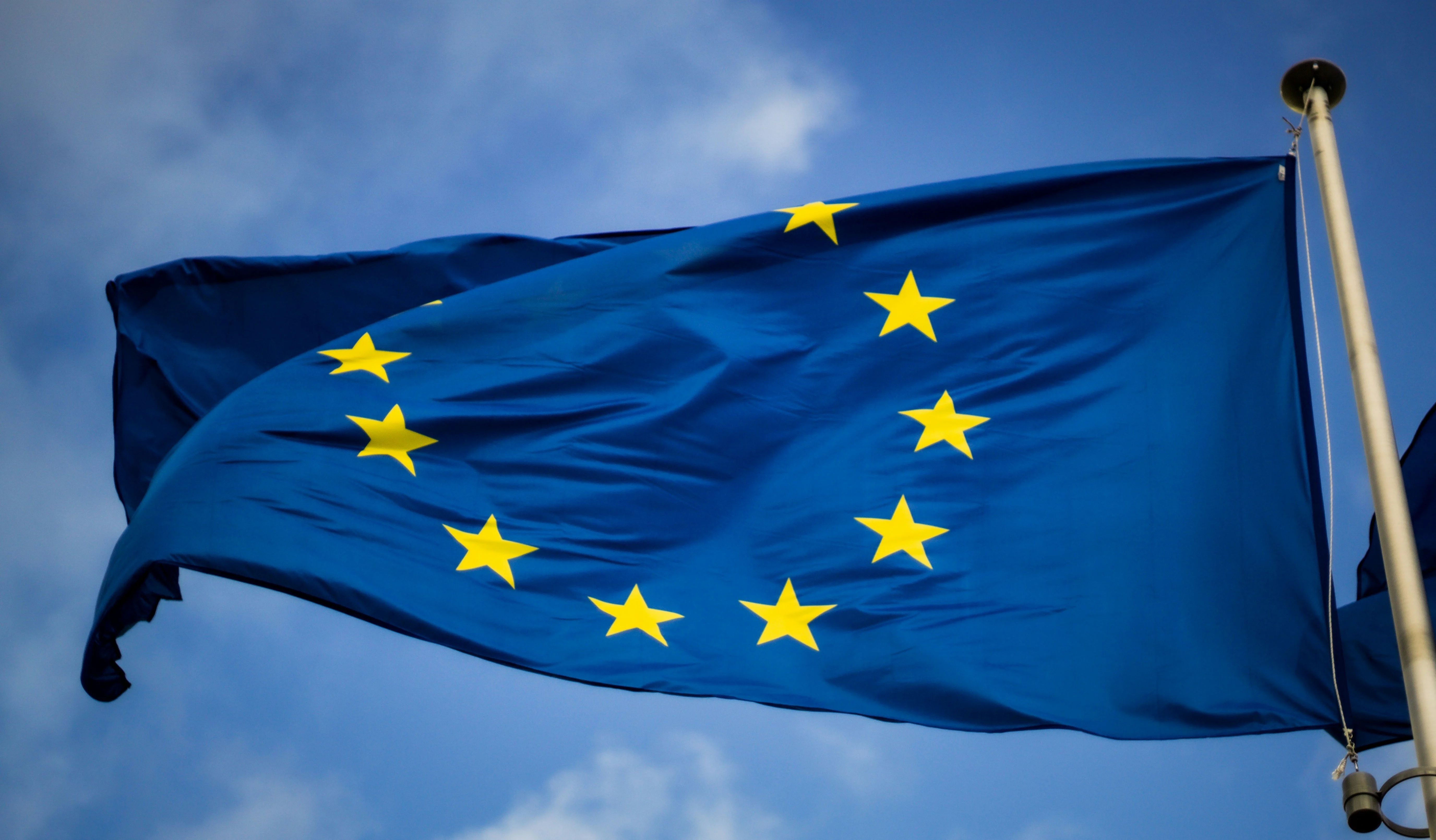 The European Union flag with yellow stars on a blue background waves against a clear sky.