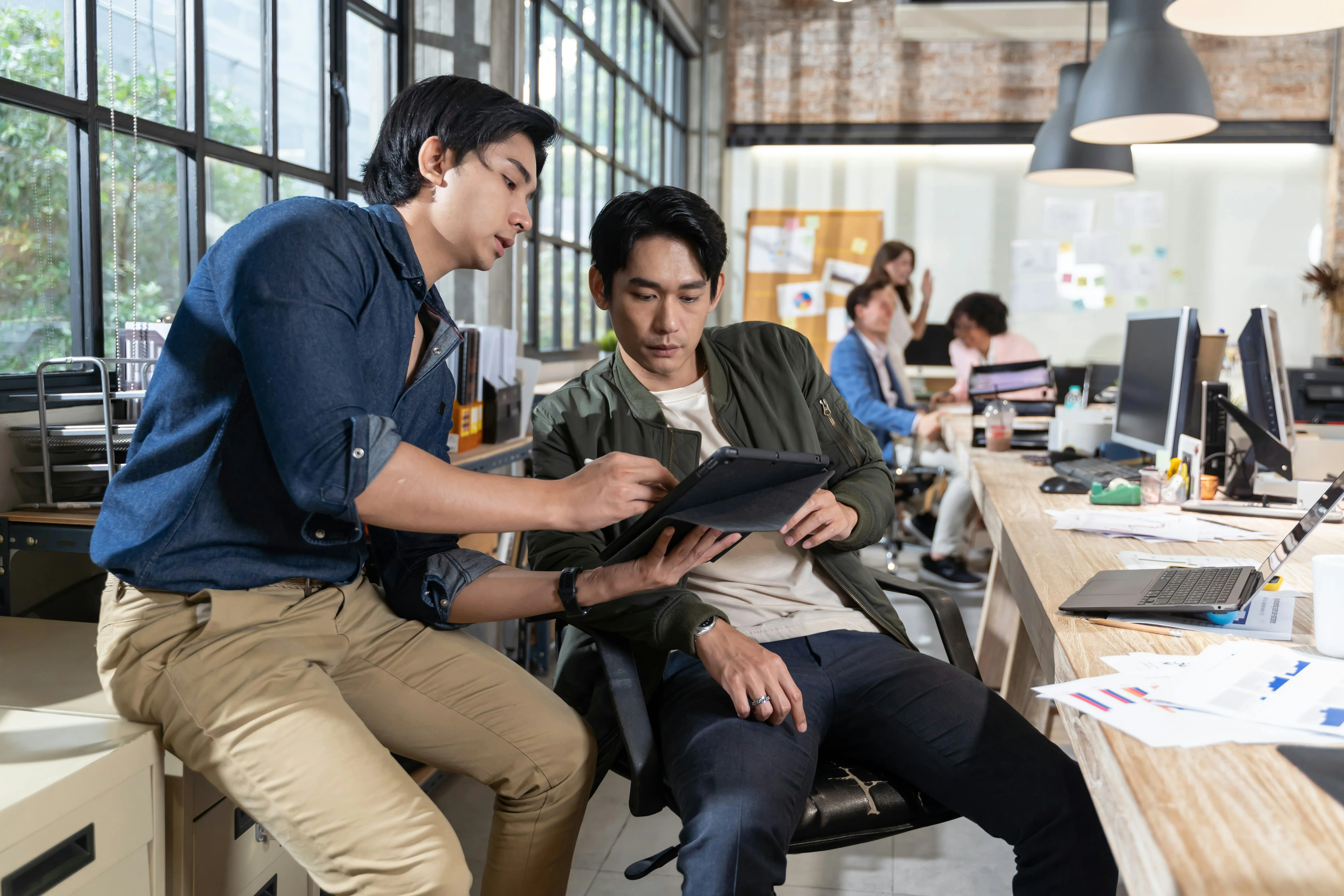 Two men discussing over a tablet in a modern office. Others work at desks in the background, with large windows and hanging lights.