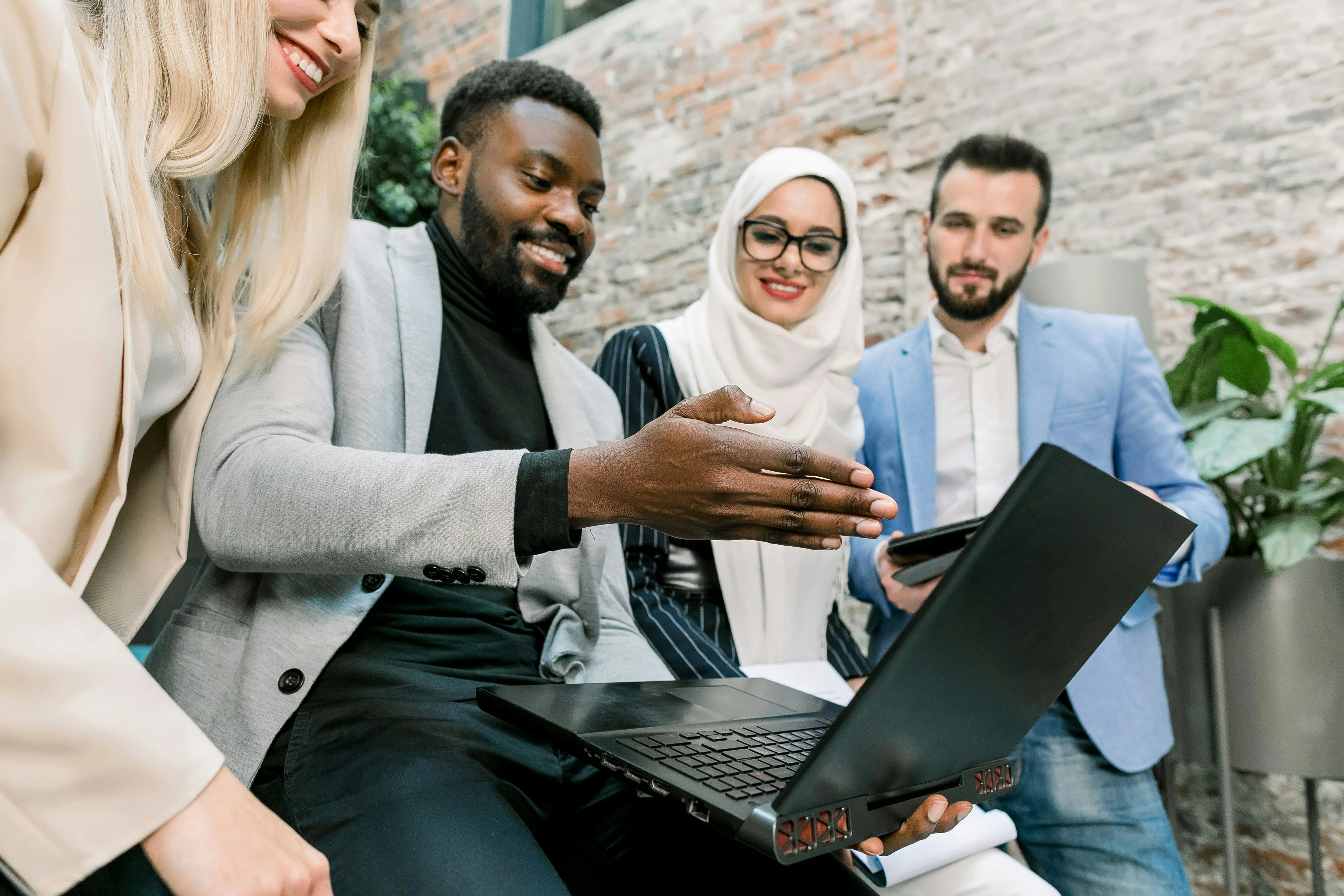 A diverse group of four people smiling and looking at a laptop, gathered in a modern office with exposed brick walls and greenery.
