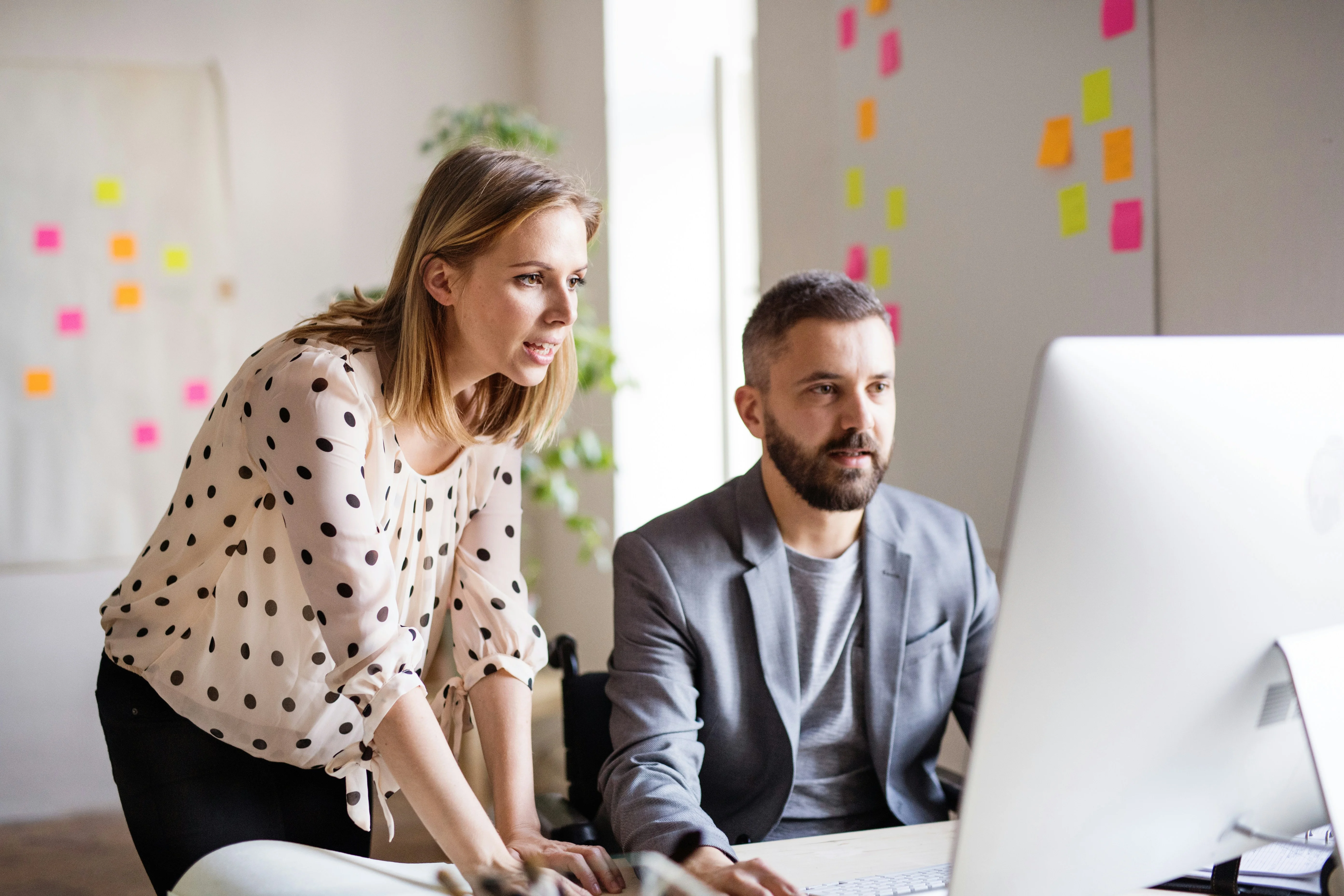 Two colleagues in a bright office, one standing and one seated, focused on a computer screen, with colorful sticky notes on the wall behind them.