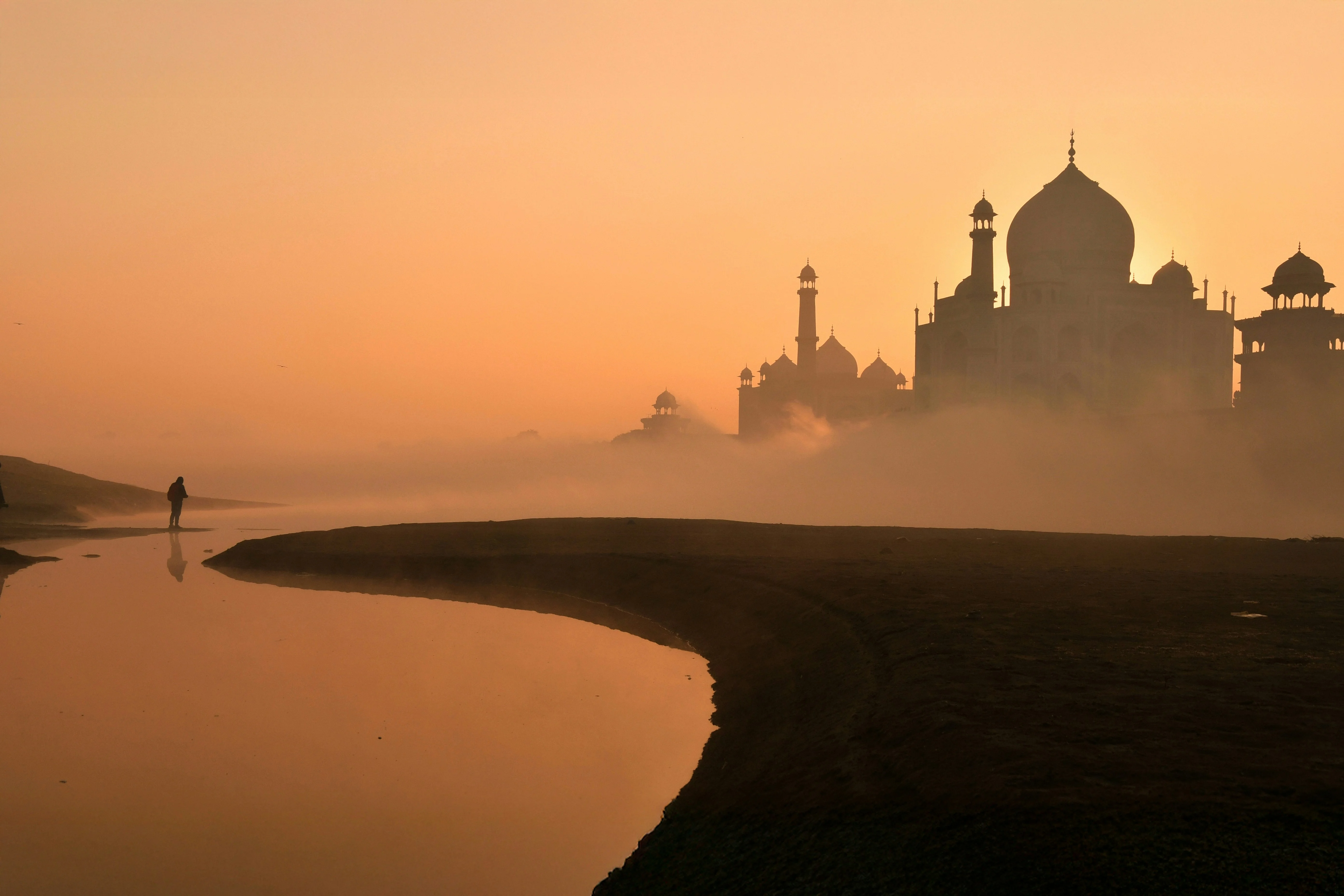 Silhouette of the Taj Mahal at sunrise, with an orange sky, mist, and a person walking near the curved riverbank.