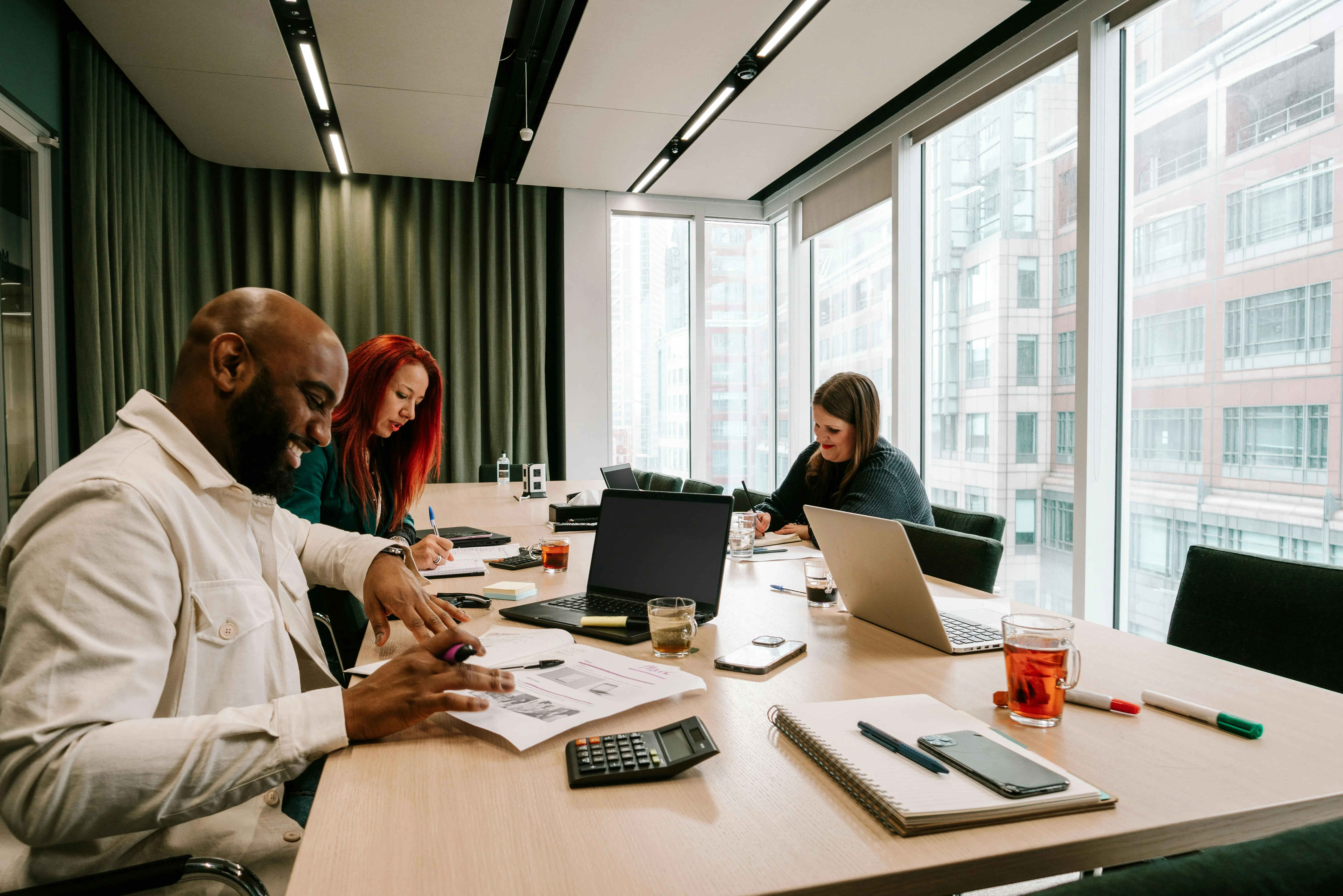Three people in a modern office meeting room, working with laptops and documents on a table, with large windows in the background.