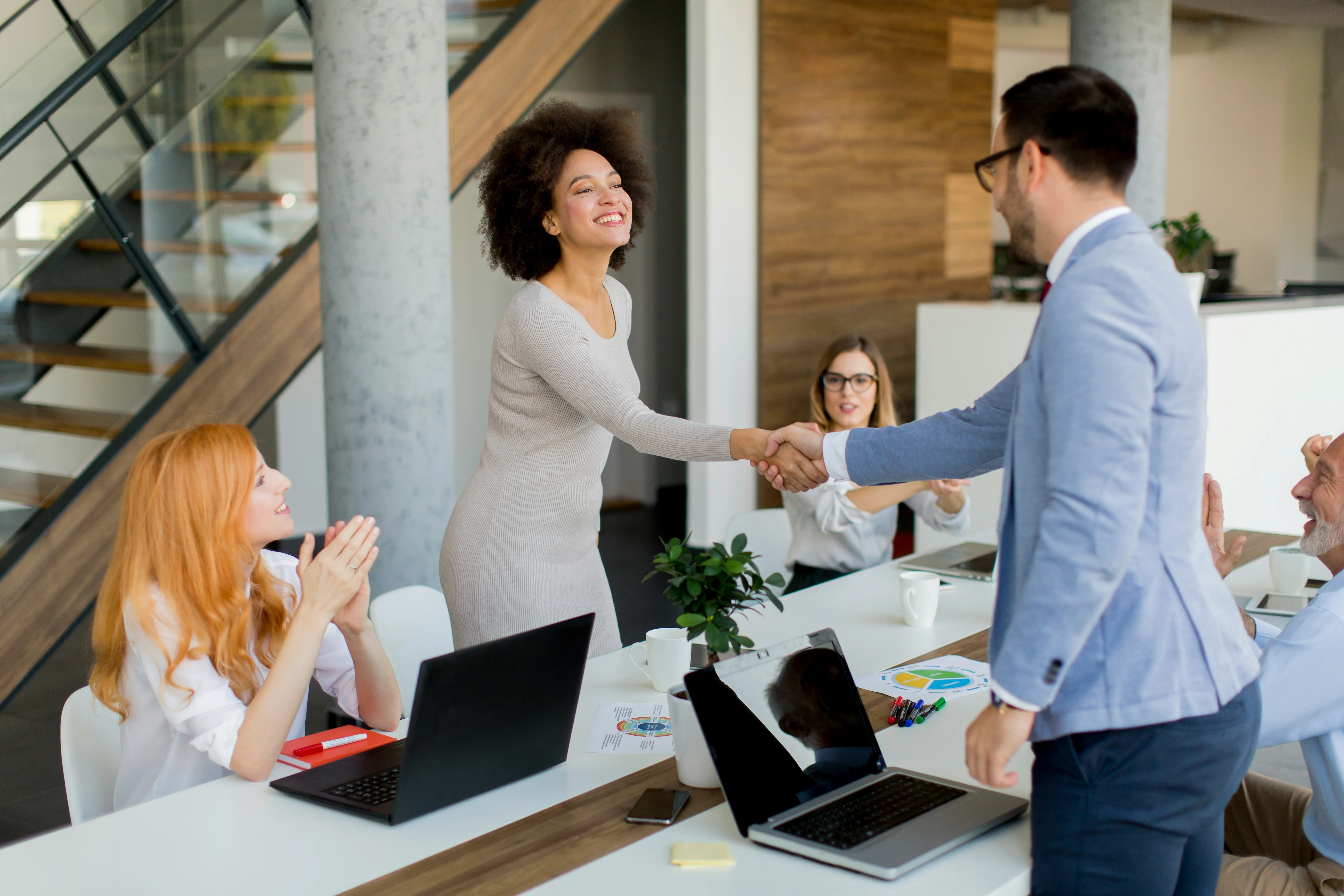 A woman stands and shakes hands with a man in a modern office, while two colleagues sit nearby, one clapping and the other smiling.