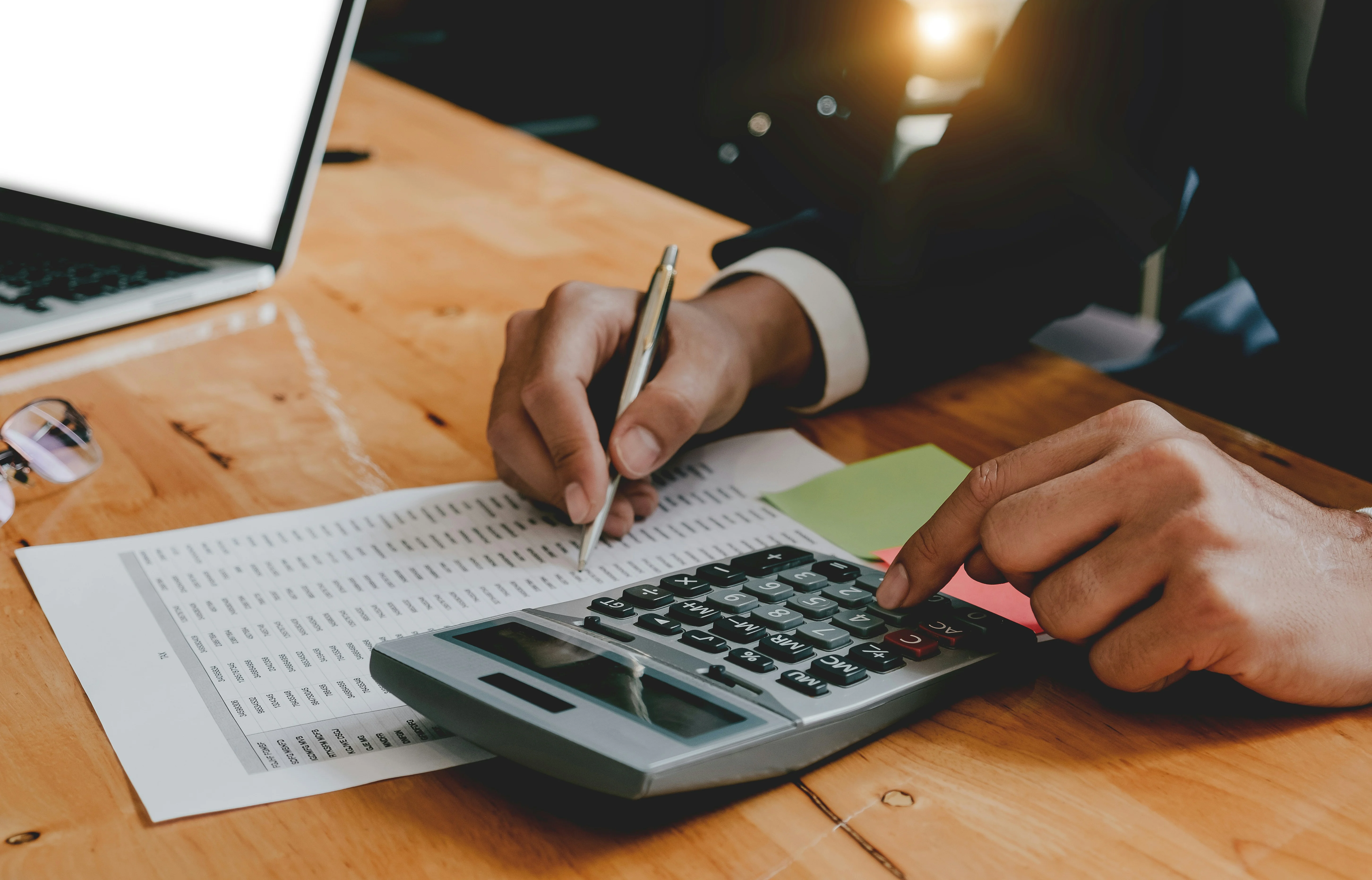 Person using a calculator and pen on a document at a wooden desk, with a laptop nearby.