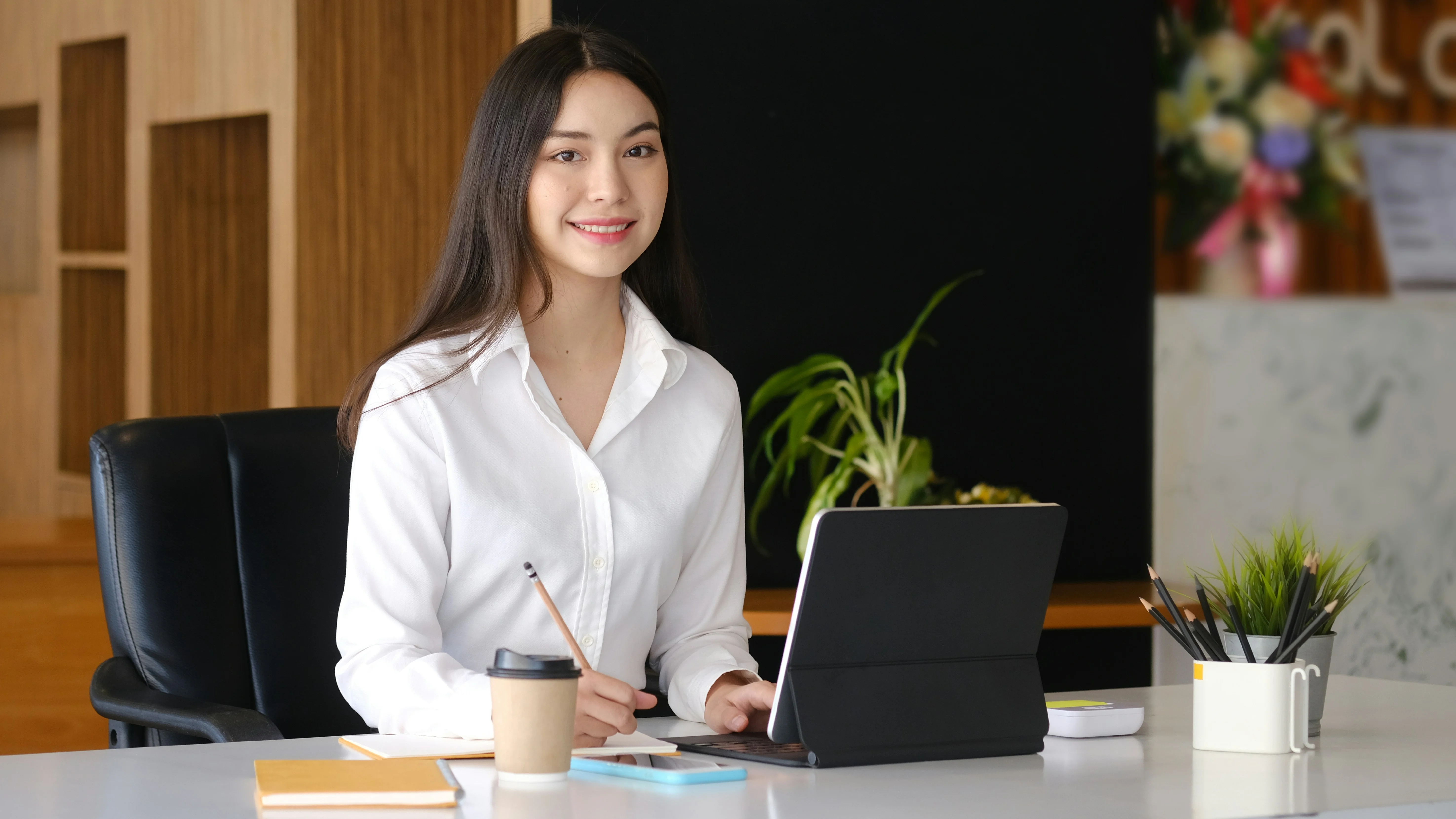 Woman in a white shirt sitting at a desk with a tablet, holding a pencil, and smiling. Office setting with plants and office supplies.