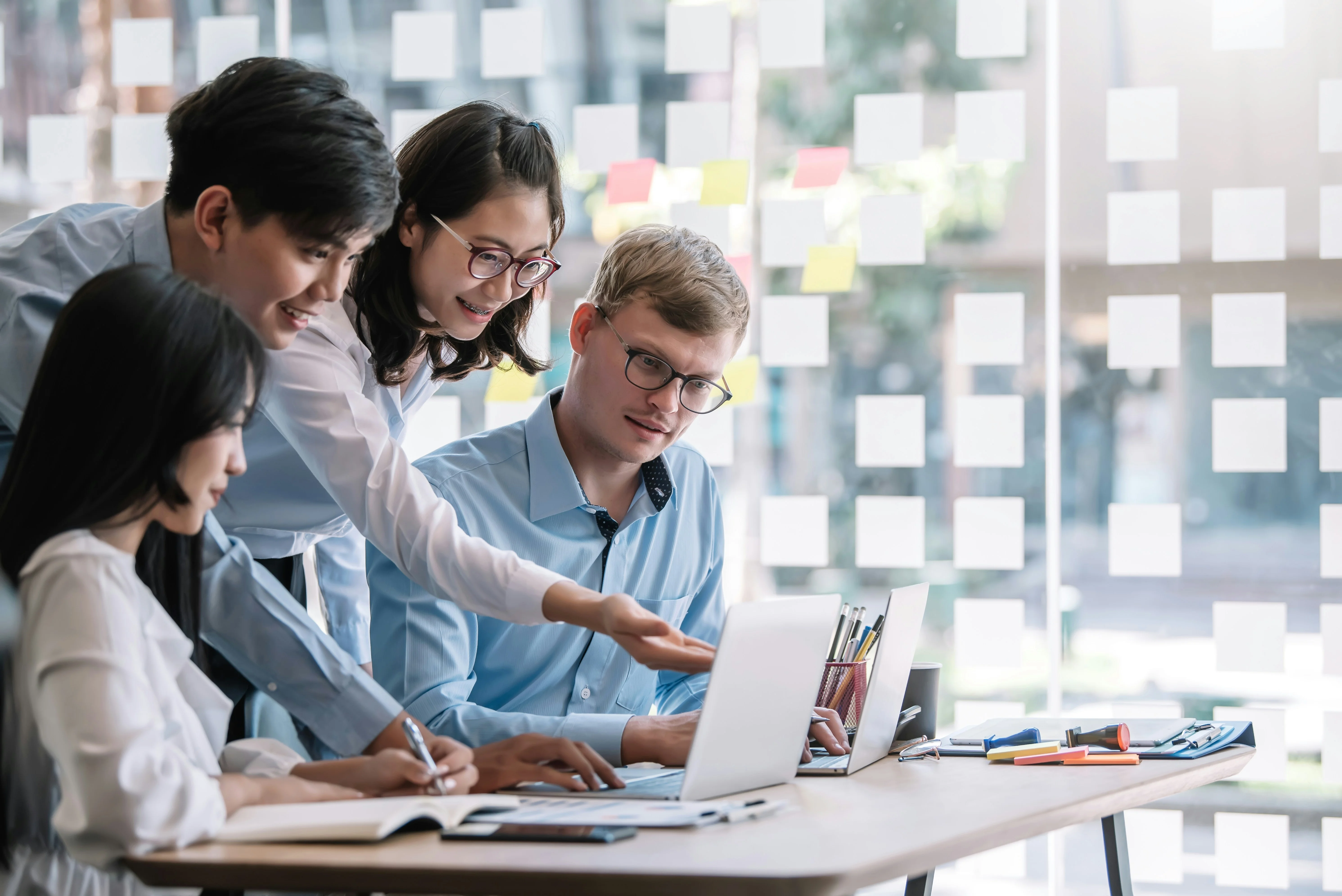 Four colleagues collaborate at a desk with laptops and notes, surrounded by a glass wall with sticky notes.