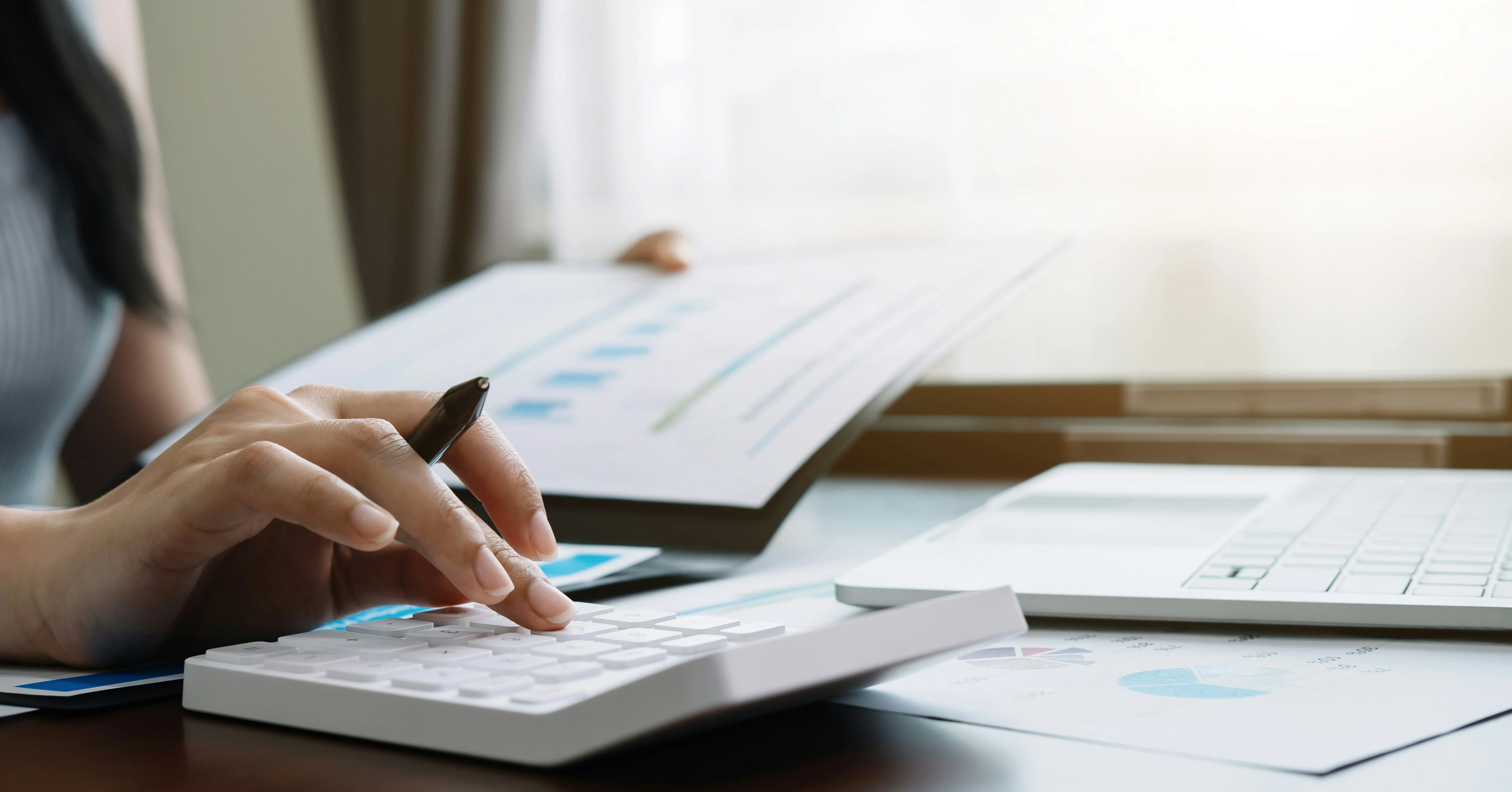 Person using a calculator and holding documents near a laptop on a desk, focused on financial calculations or data analysis.
