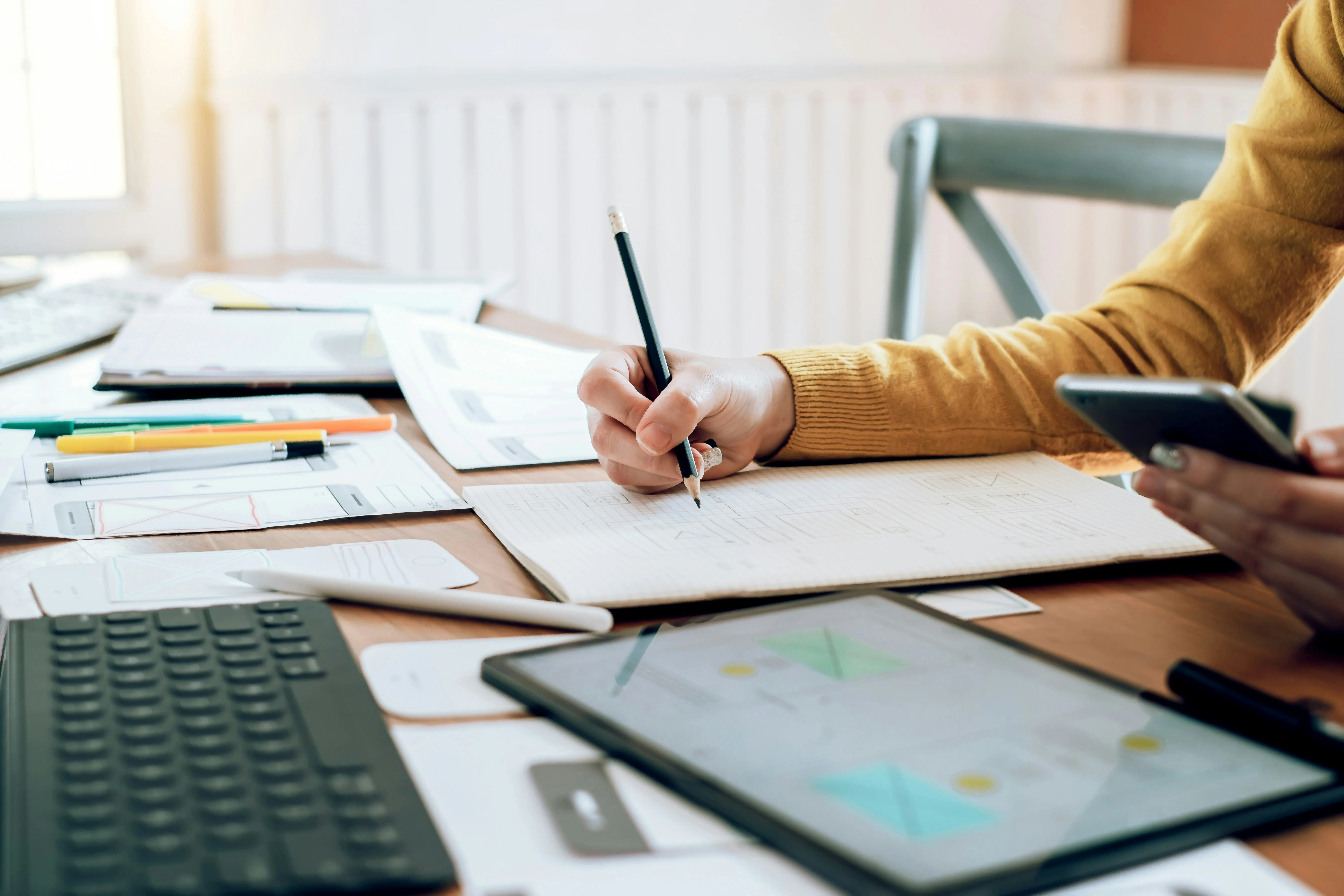Person in a yellow sweater writing in a notebook while holding a smartphone, surrounded by papers, a tablet, and a keyboard on a desk.