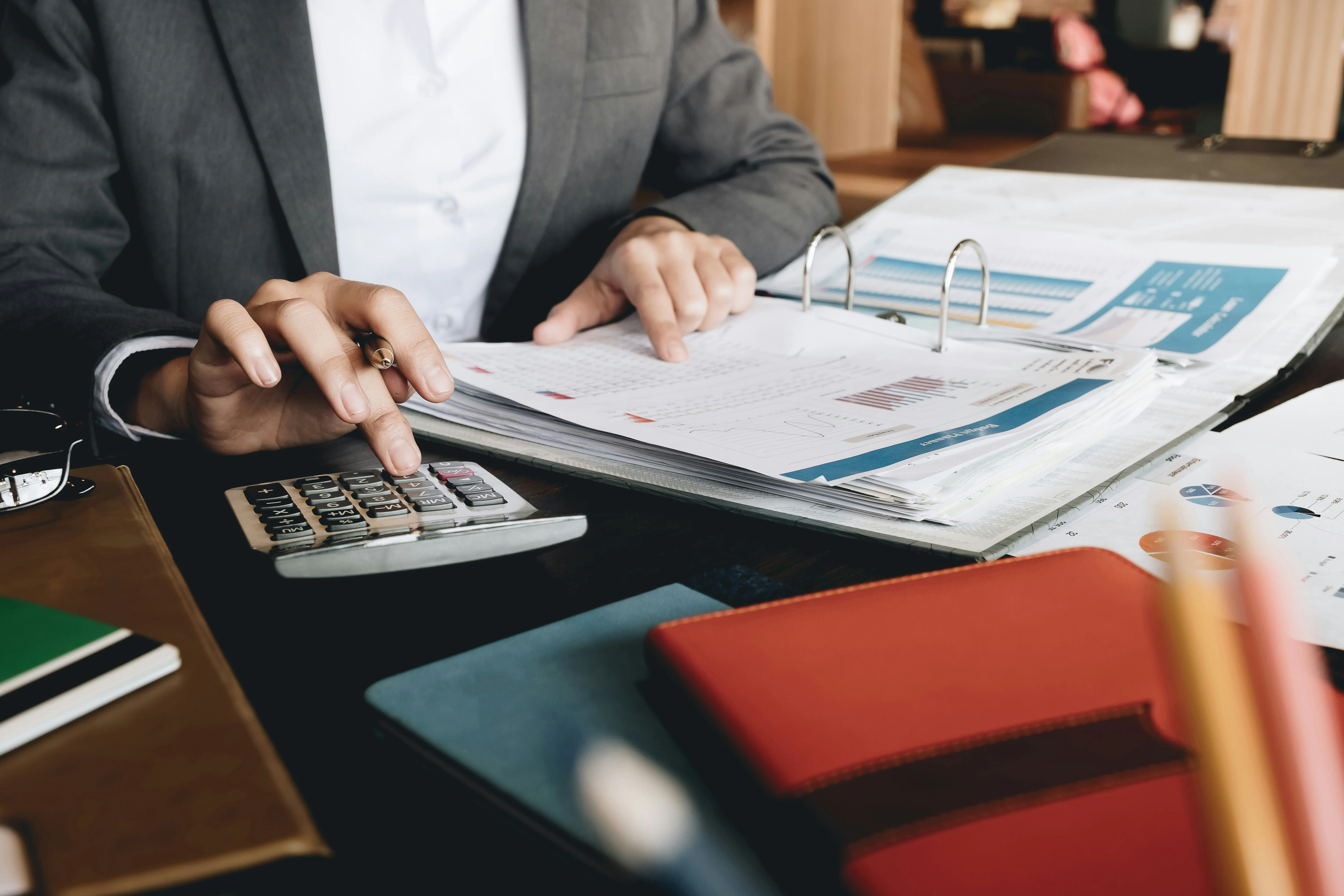 Person in business suit using a calculator and reviewing financial reports in a binder on a cluttered desk with notebooks and charts.