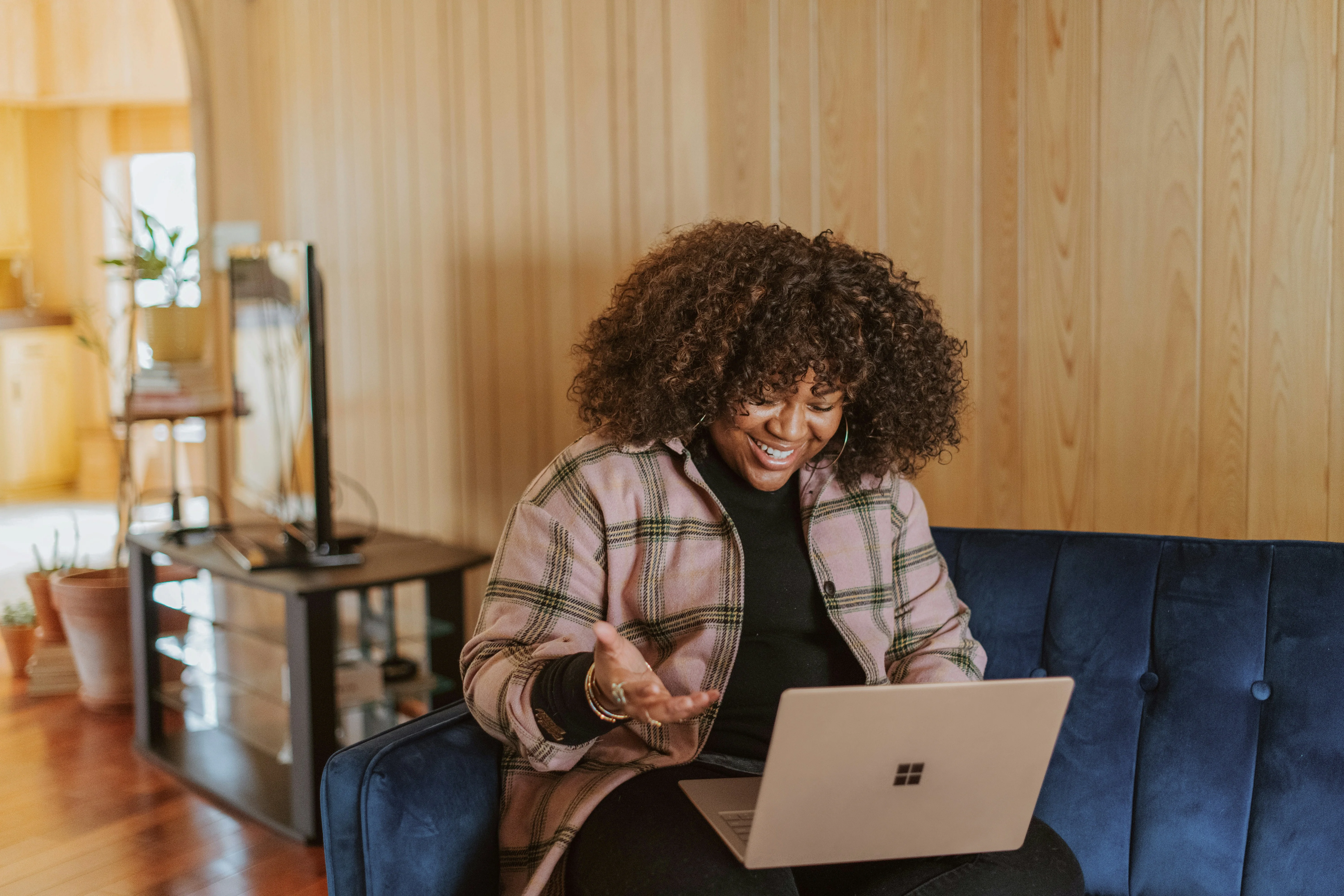 Person with curly hair, wearing a plaid jacket, smiling and gesturing while using a laptop on a blue sofa in a wood-paneled room.