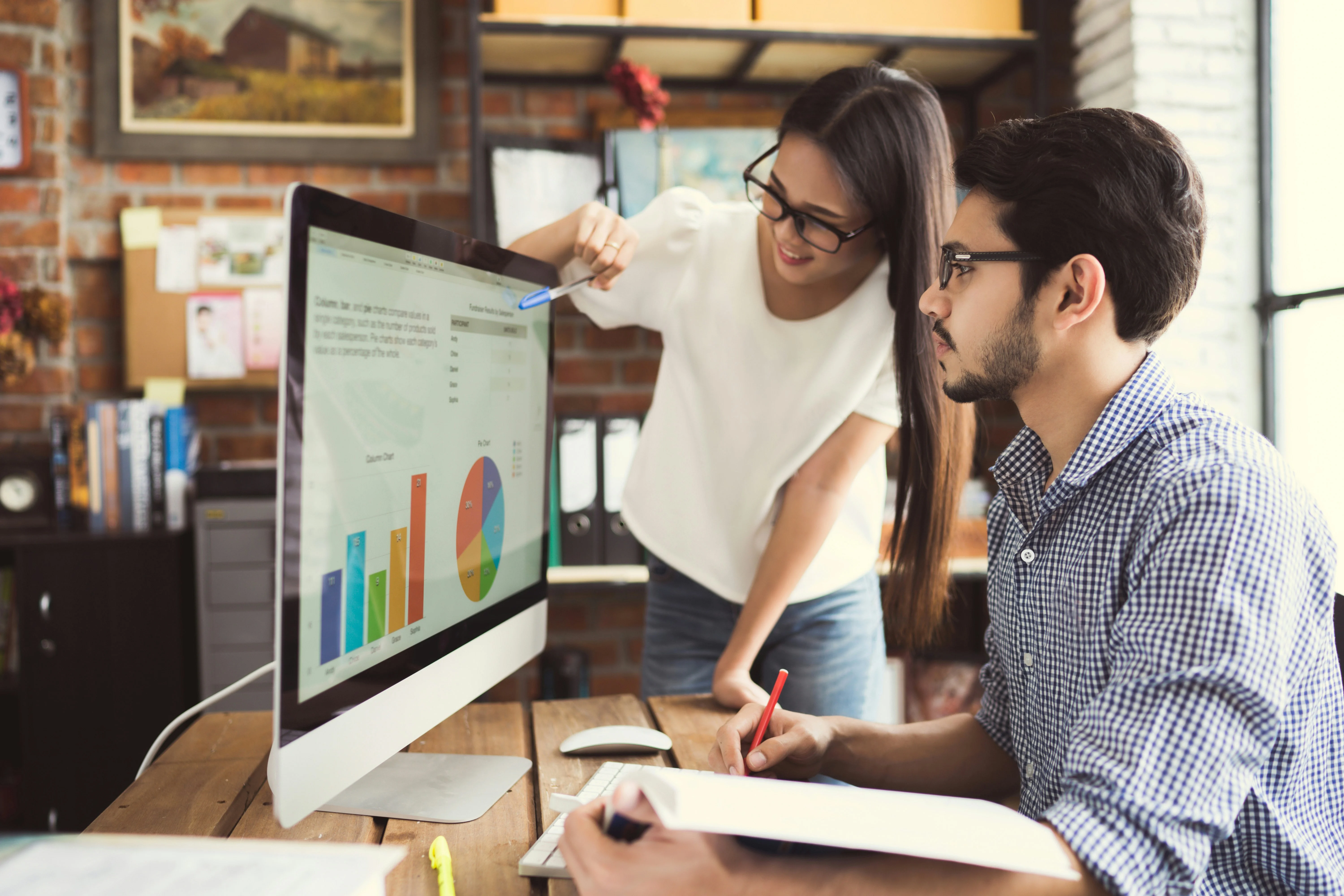 Two coworkers analyze graphs on a computer in an office. One points at the screen with a pen, while the other takes notes.