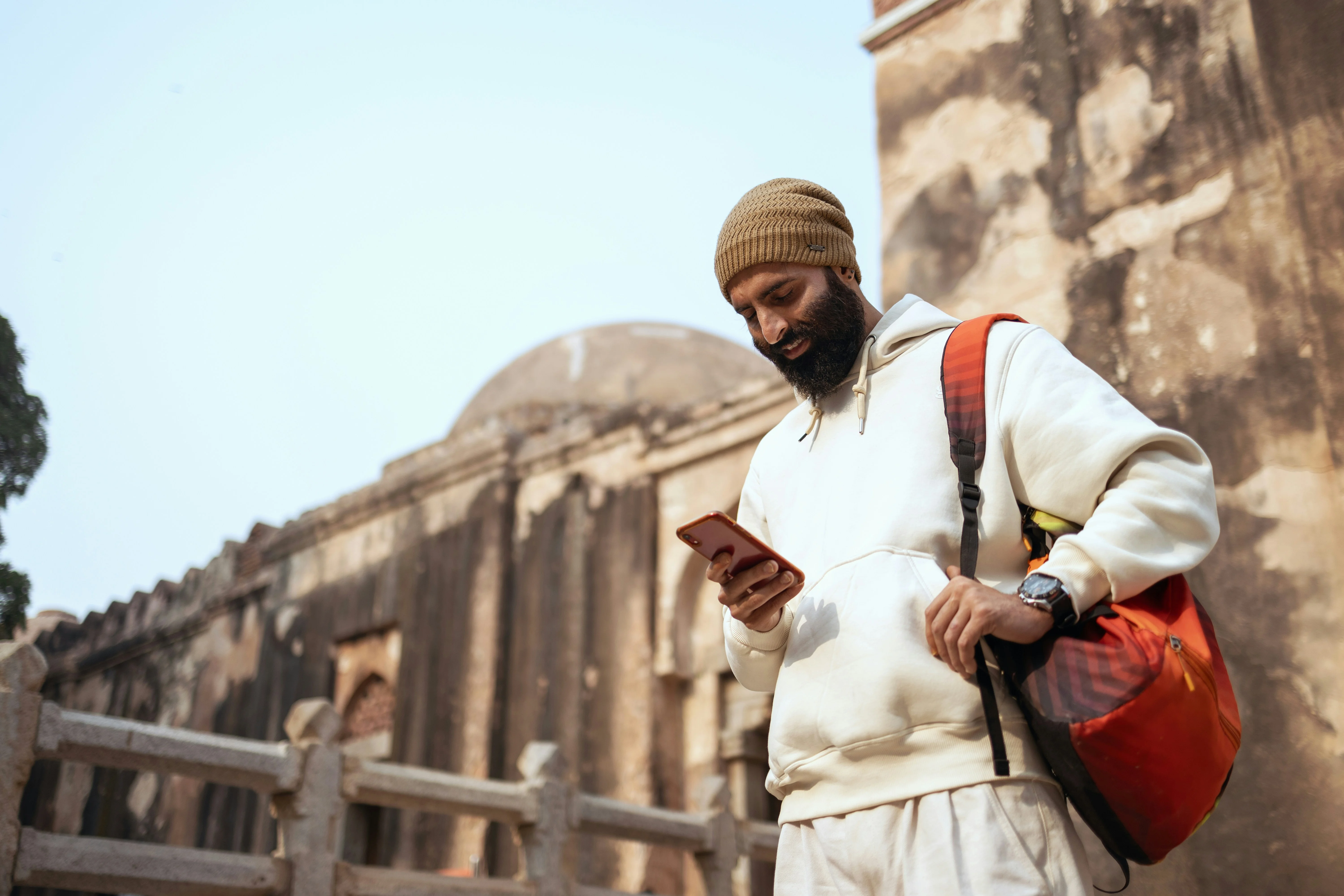 Man in a white hoodie and beanie checks his phone while standing near an ancient stone building with a backpack.