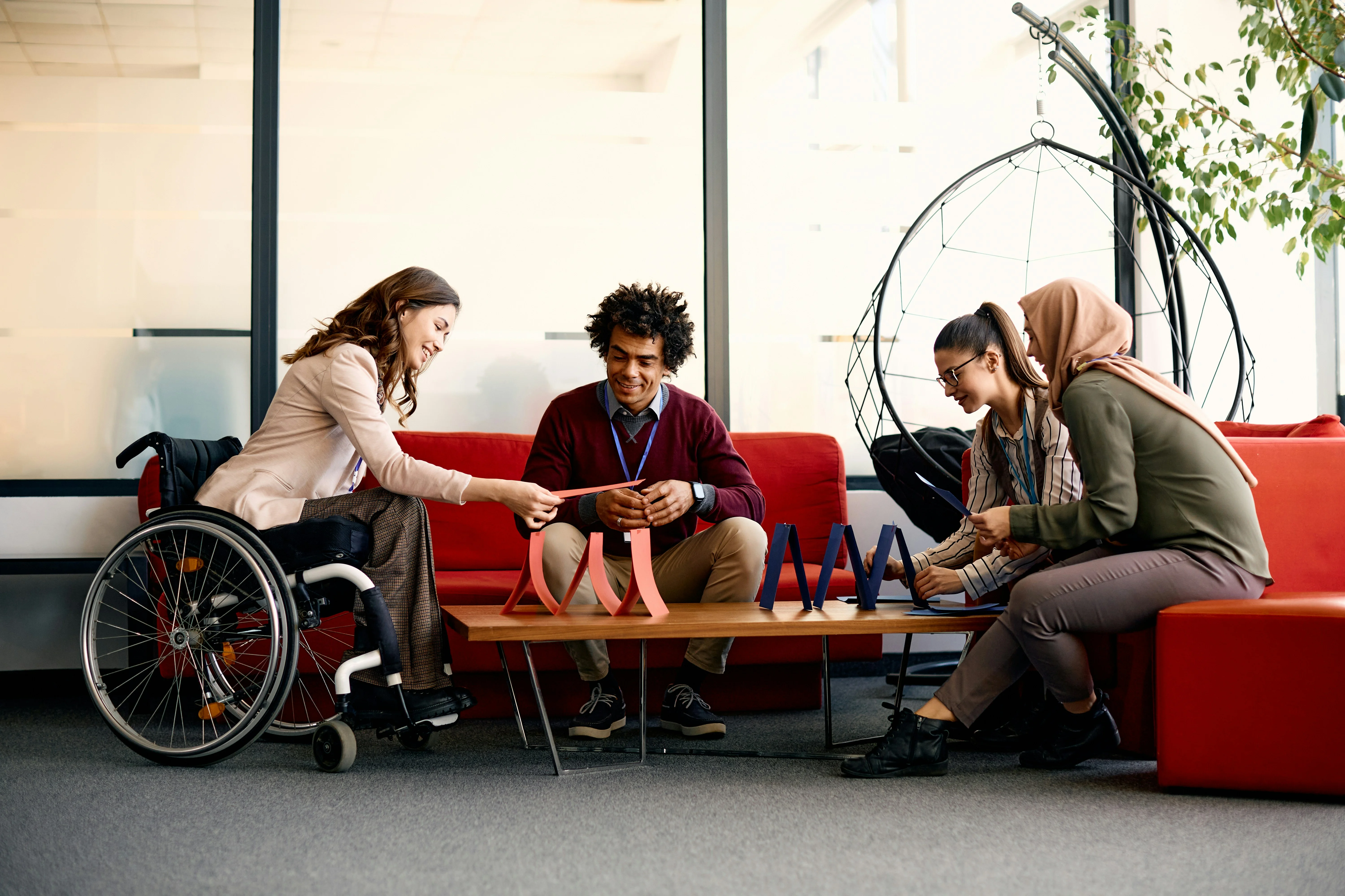 Four people, including one in a wheelchair, collaborating around a table with colorful objects in a modern office setting.