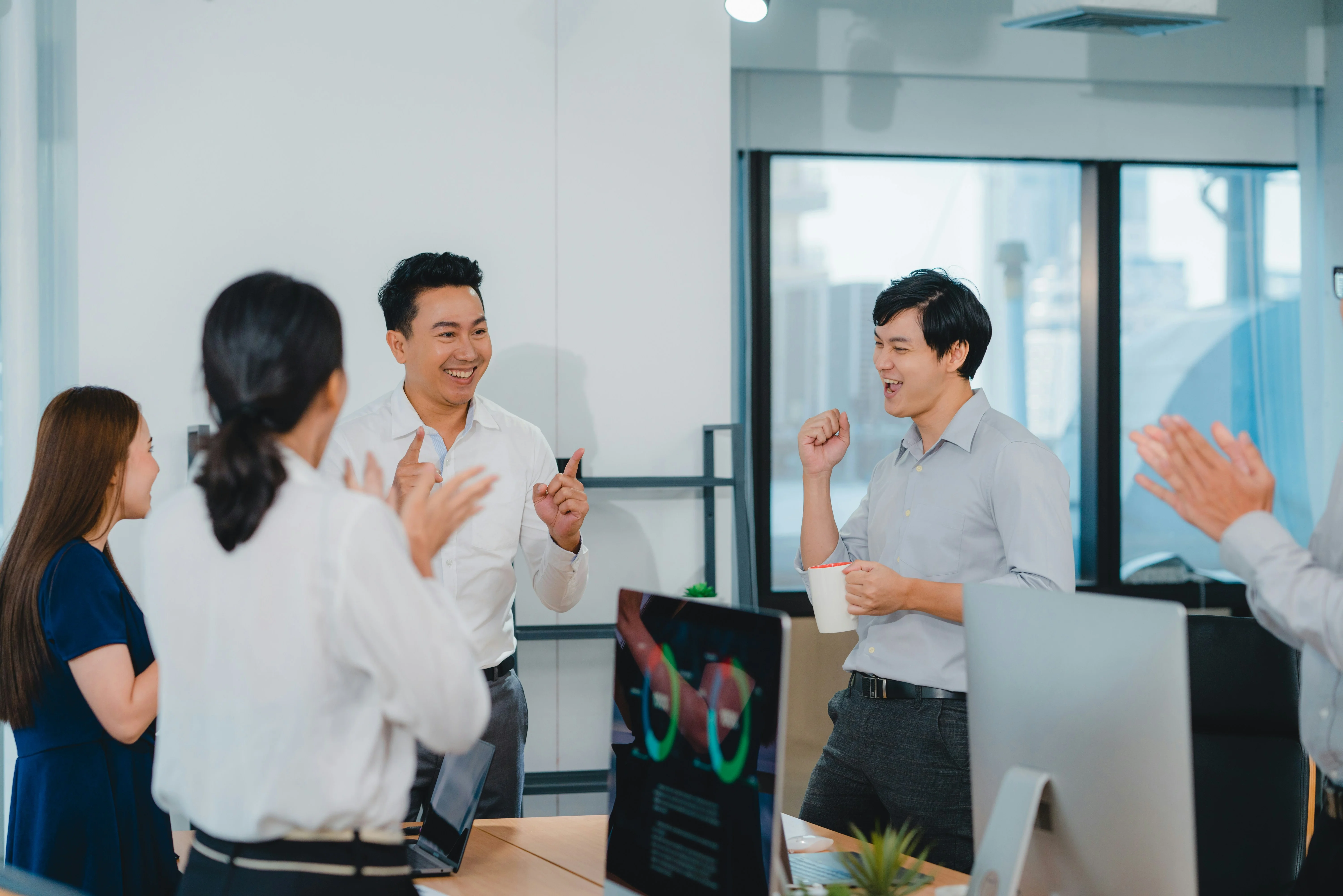Coworkers in an office celebrate a success, with two men smiling and gesturing, while others clap. Laptops and office decor are visible.