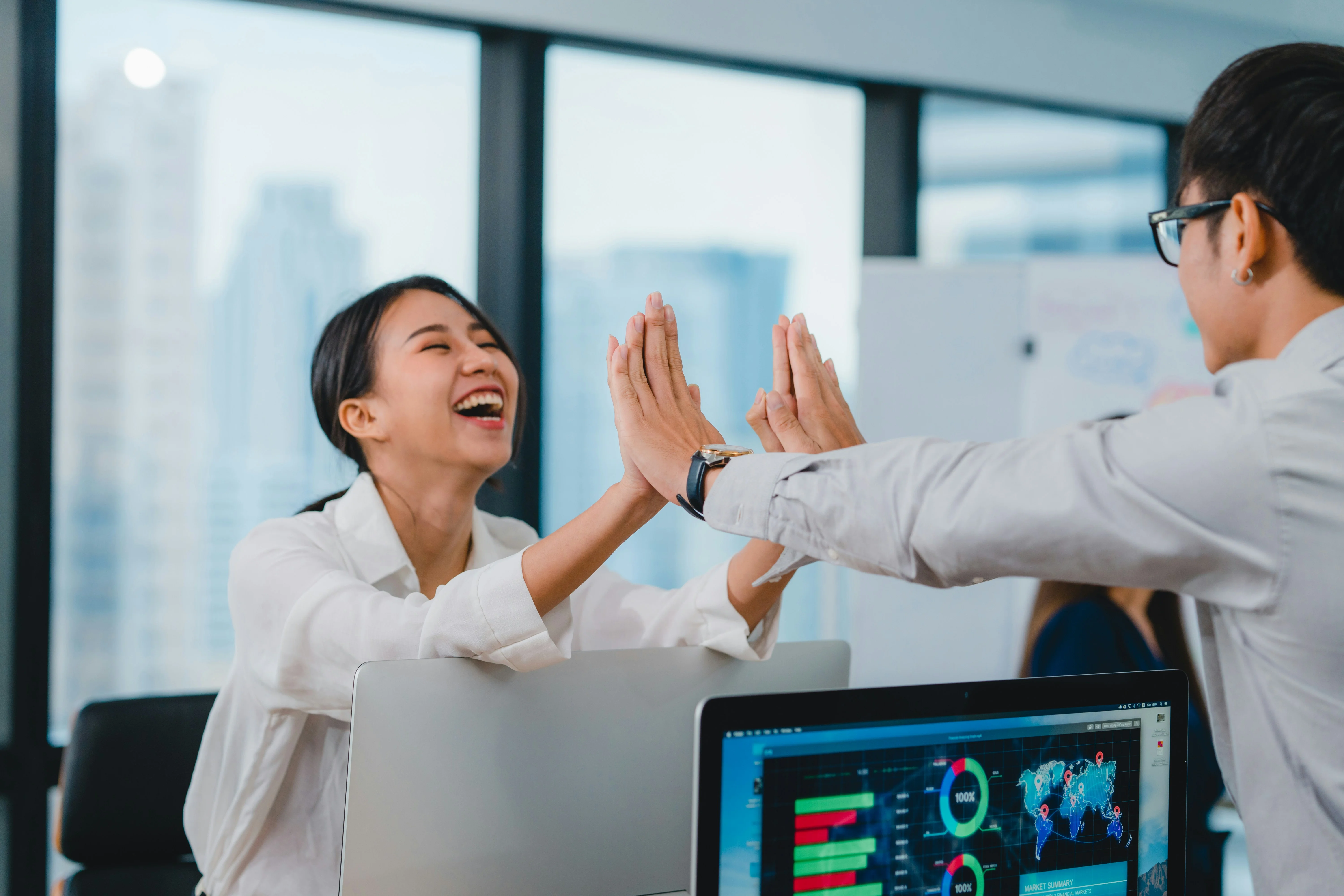 Two colleagues high-fiving and smiling in a bright office, with a computer displaying charts in the foreground.