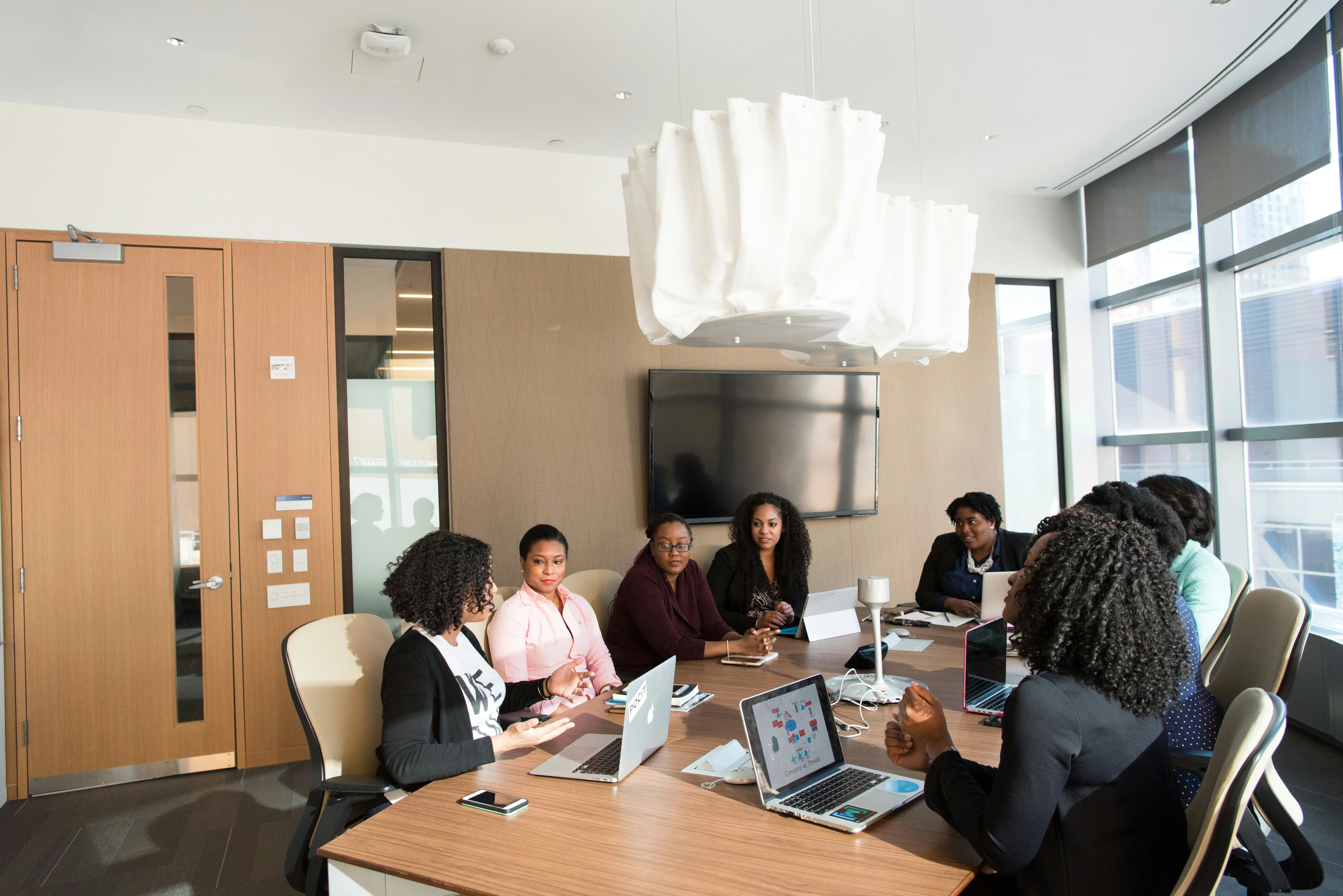 A diverse group of people in a conference room, sitting around a table with laptops and notes, engaged in a meeting.