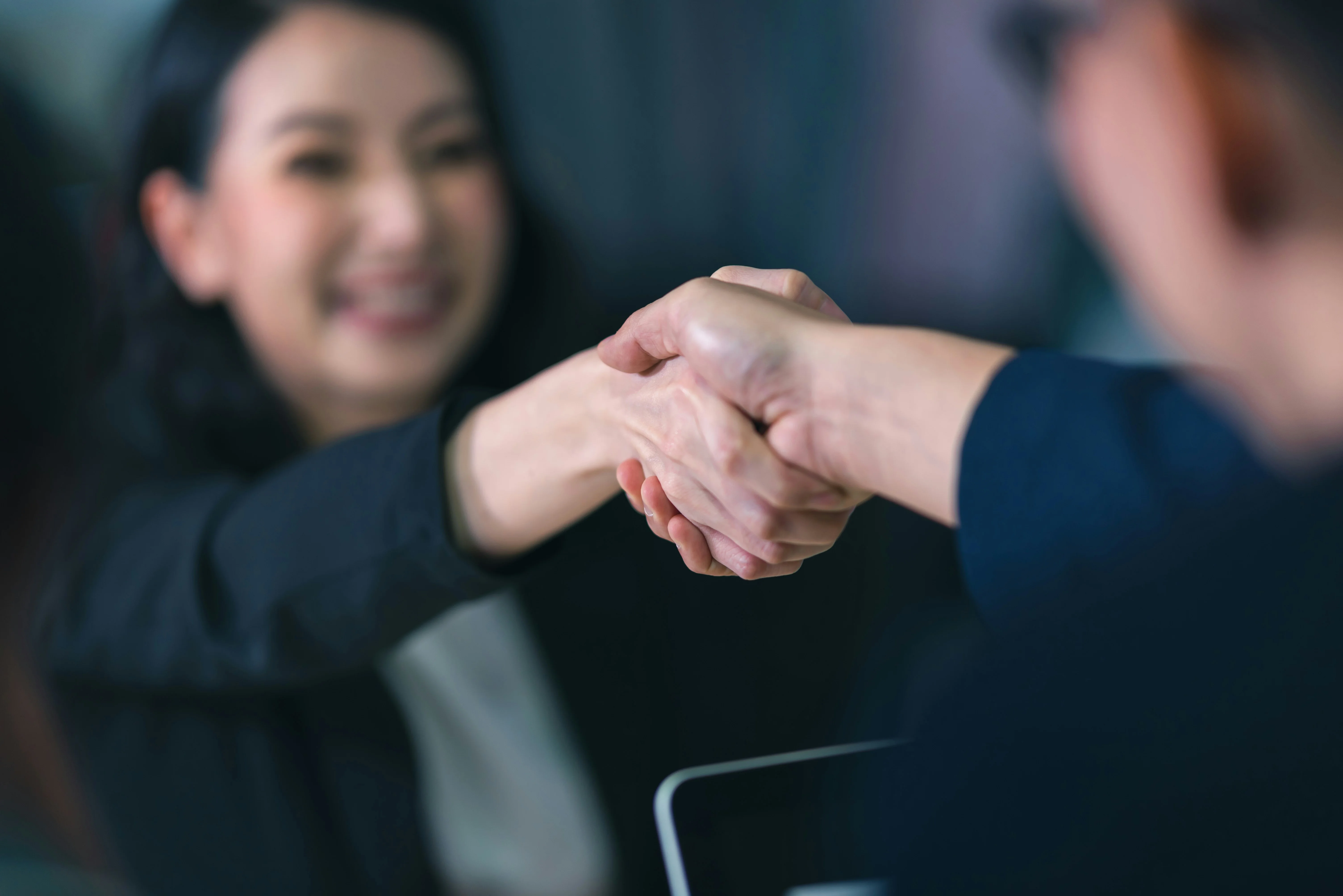 Two people shaking hands in a professional setting, with one woman smiling in the background, suggesting agreement or partnership.